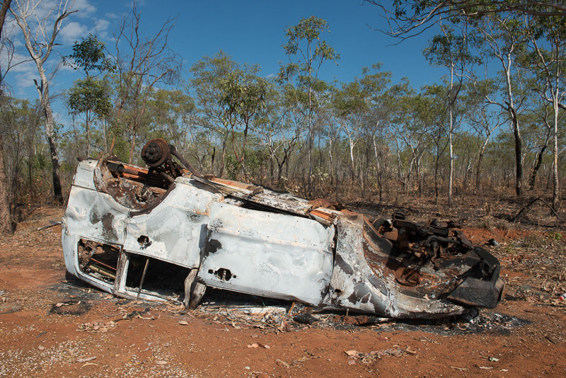 An overturned, burnt-out, rusting car on the side of a road in the Australian bush.