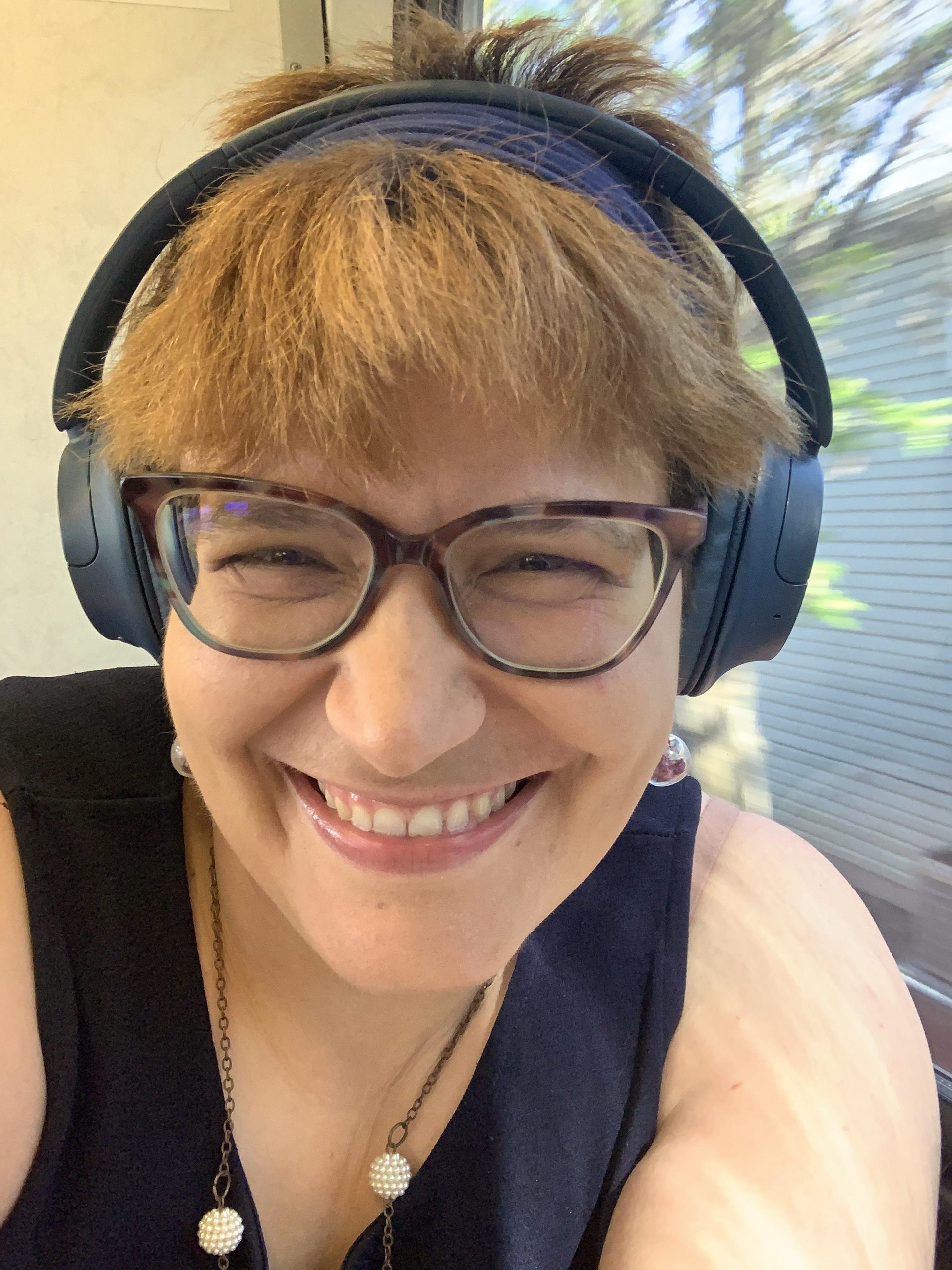 A woman grins widely as she takes a camera selfie. She is wearing noise-cancelling earphones, cat-eye glasses, a tank top, and bauble earrings. 