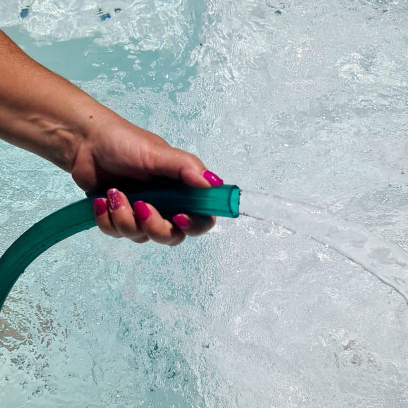 Mano sosteniendo un tubo flexible de PVC verde mientras fluye agua, sobre el fondo de una pileta o superficie acuática.