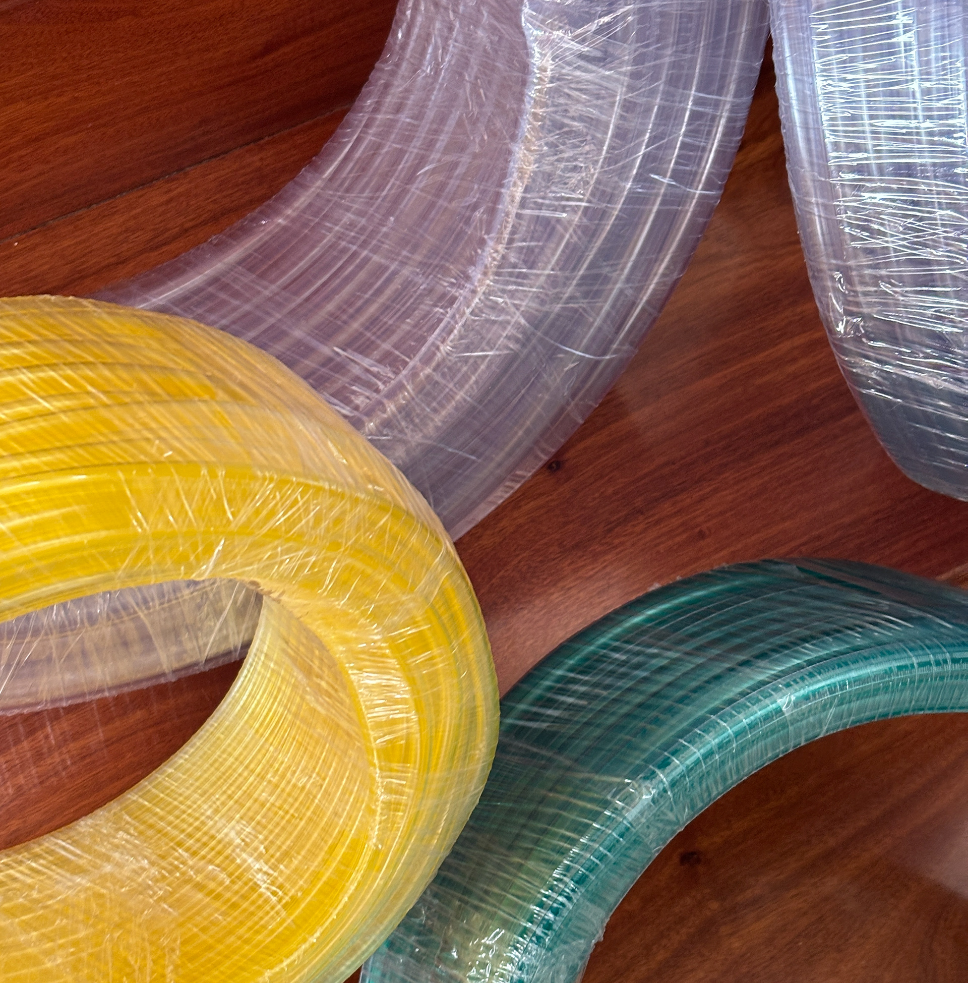 Bobinas de tubos flexibles de PVC virgen en colores transparente, amarillo y verde, envueltos en film plástico, sobre superficie de madera.