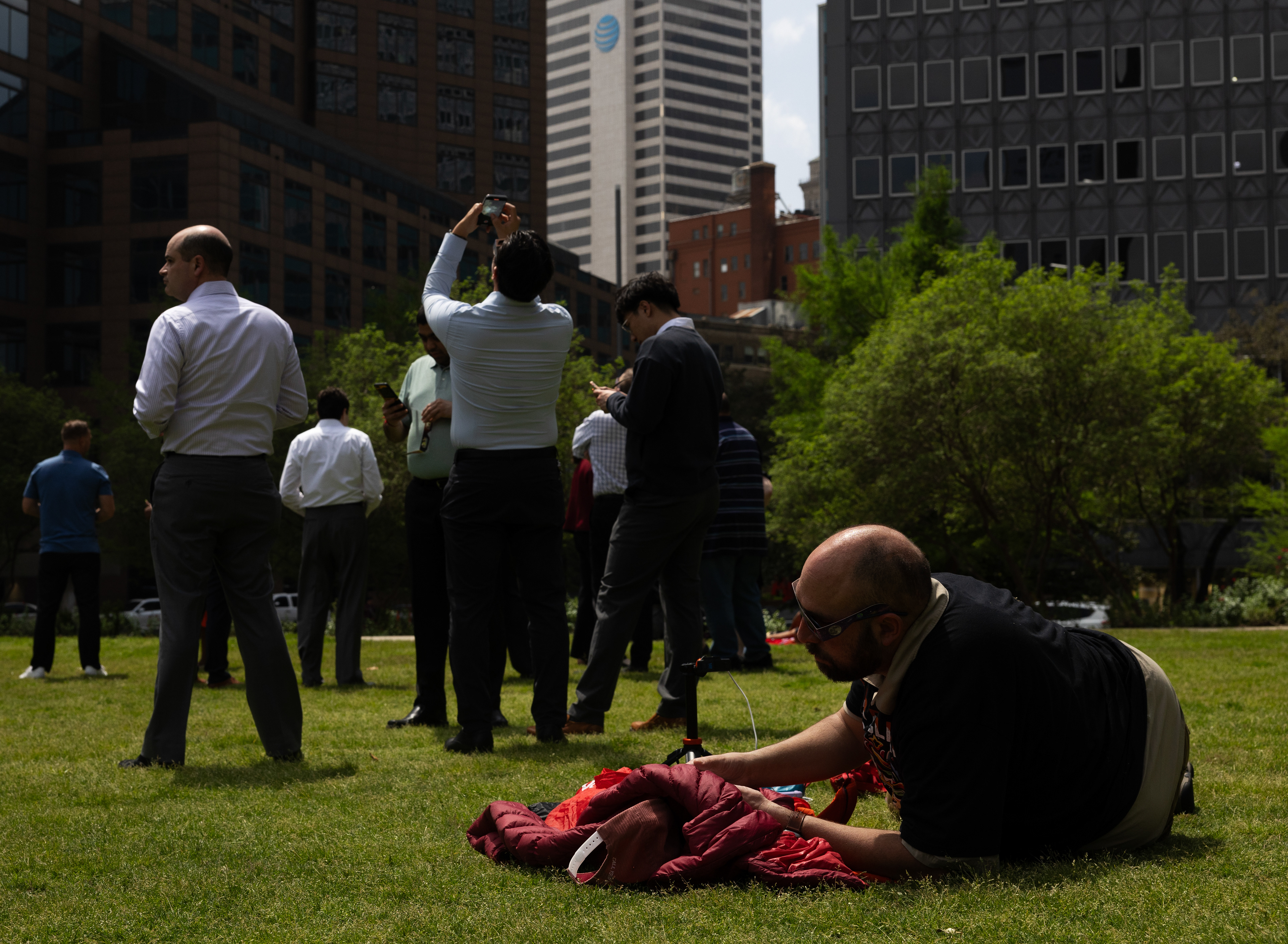 A man lays in the grass geared up to enjoy nature while watching the solar eclipse while business men come out of their skyscraper offices to experience totality outside. 