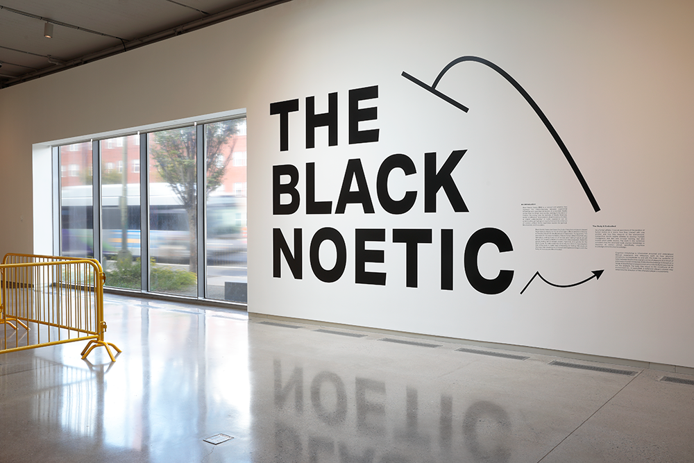 In large black text, the words "The Black Noetic" are printed on the white gallery walls.