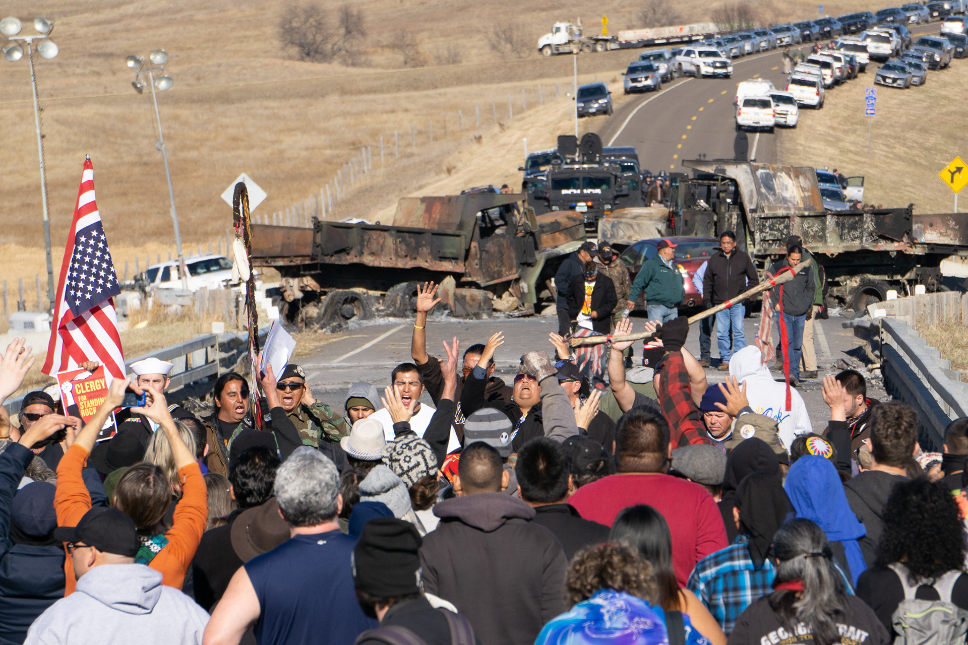 Water protectors lead a chant on Backwater Bridge near Cannon Ball, North Dakota. The burned out remains of trucks and a long line of state and federal law enforcement vehicles block the bridge.