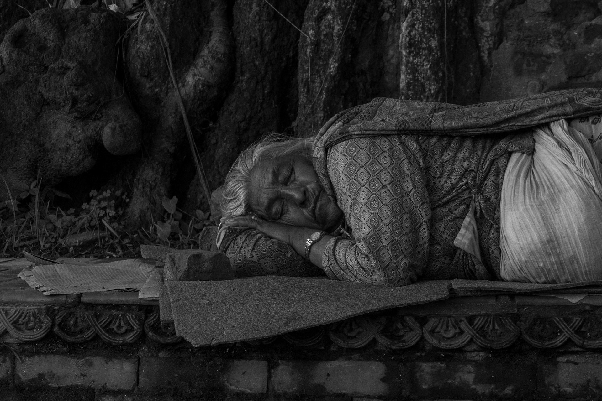 An old woman napping during lunch. She is by an old and lush tree, with her bags near her. Unbothered, her connection to nature is a strong one, something we dont see everyday. .