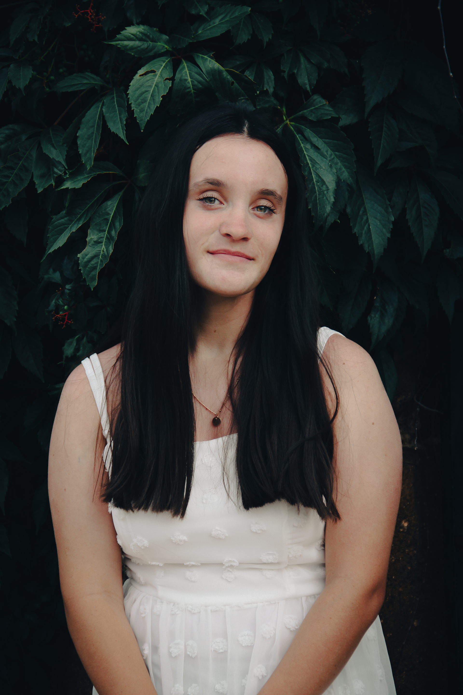 Natural light senior portrait of a young woman with long dark hair and a soft expression, wearing a white dress and standing against a dark green leafy background.