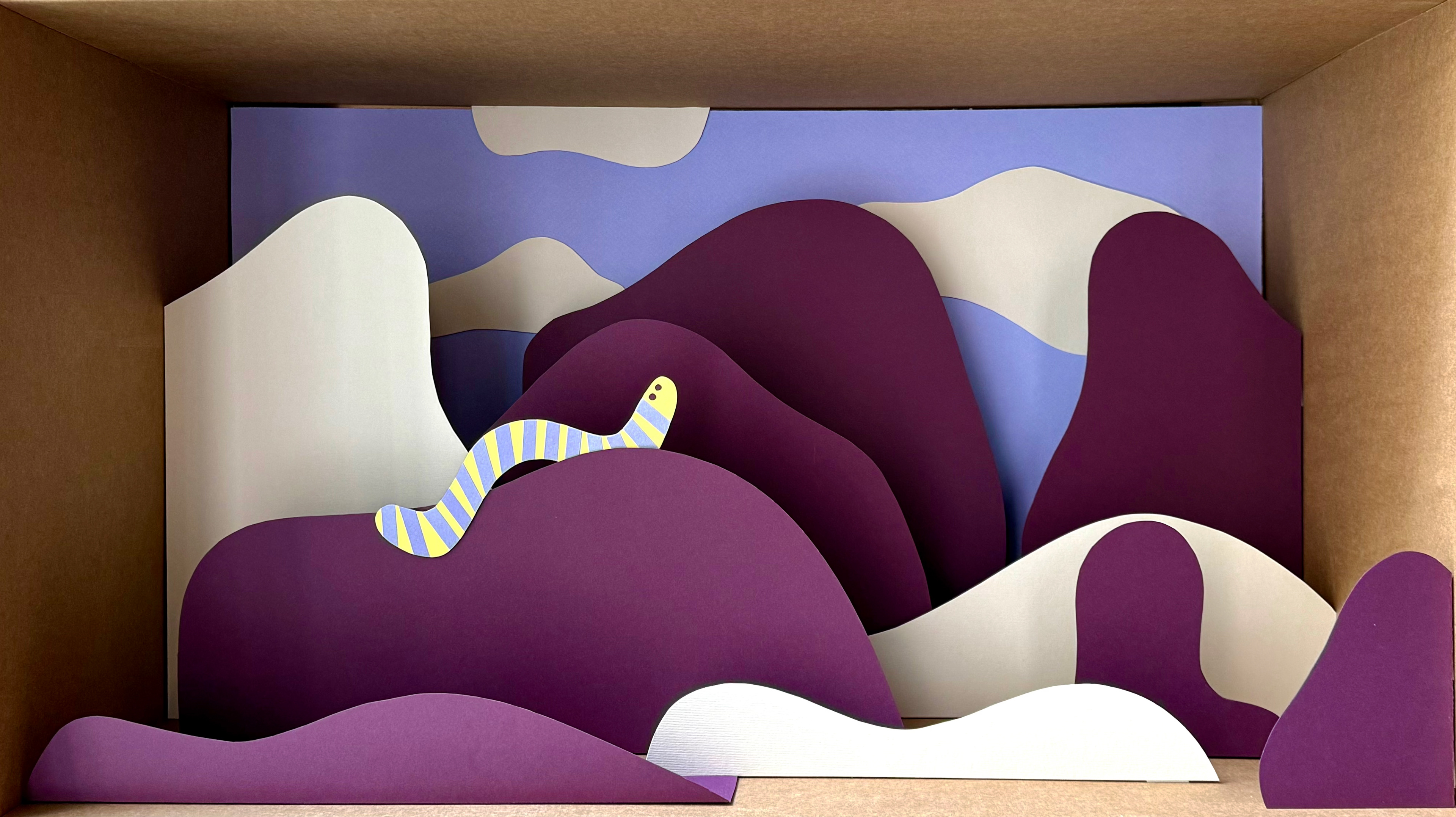 3D paper cut artwork by Helga Aversa for Illustri Hub, featuring a worm hiding in dunes with purple, yellow, and blue cardboard