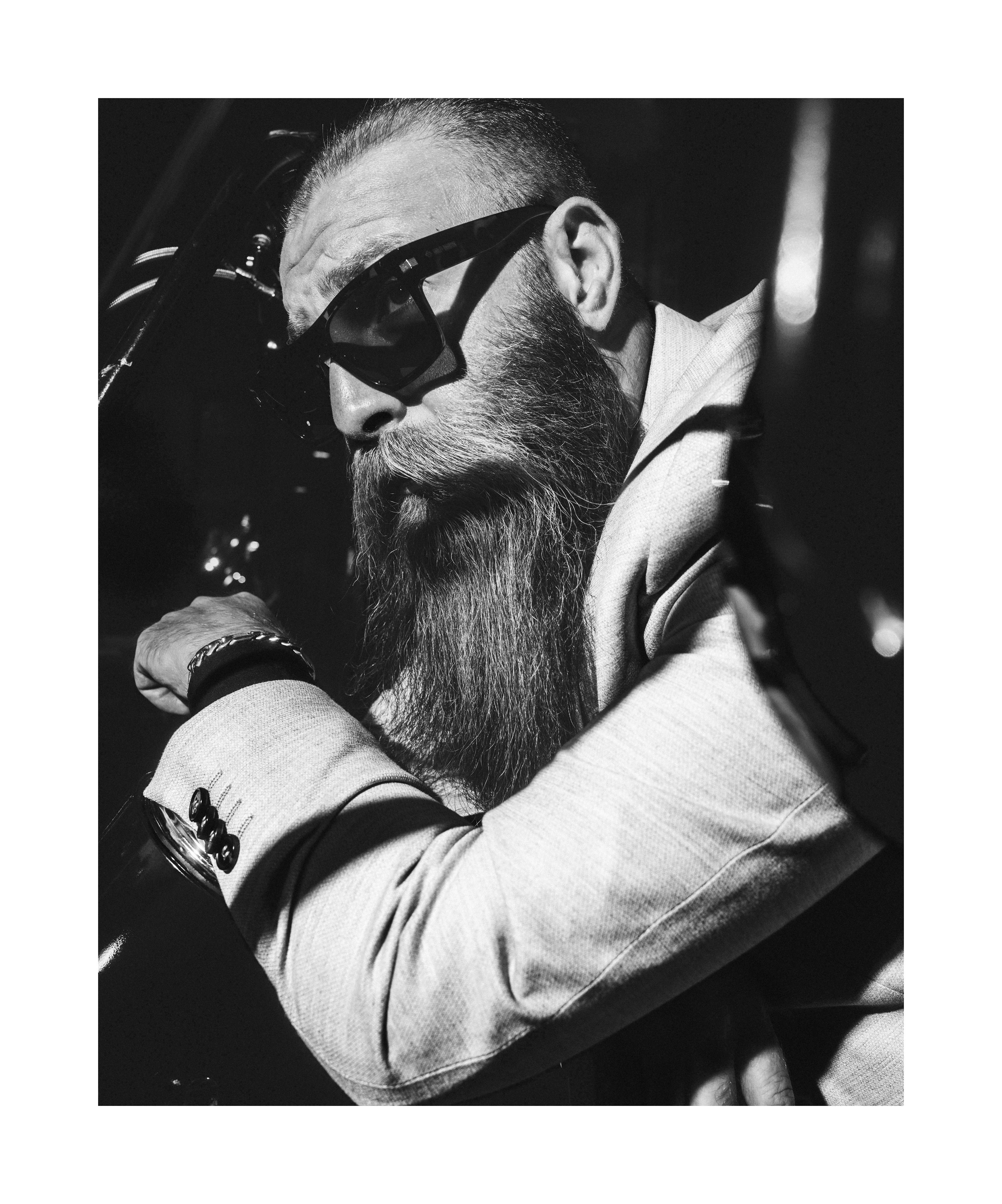 Black and white close-up portrait of a bearded man in sunglasses leaning on a motorcycle.