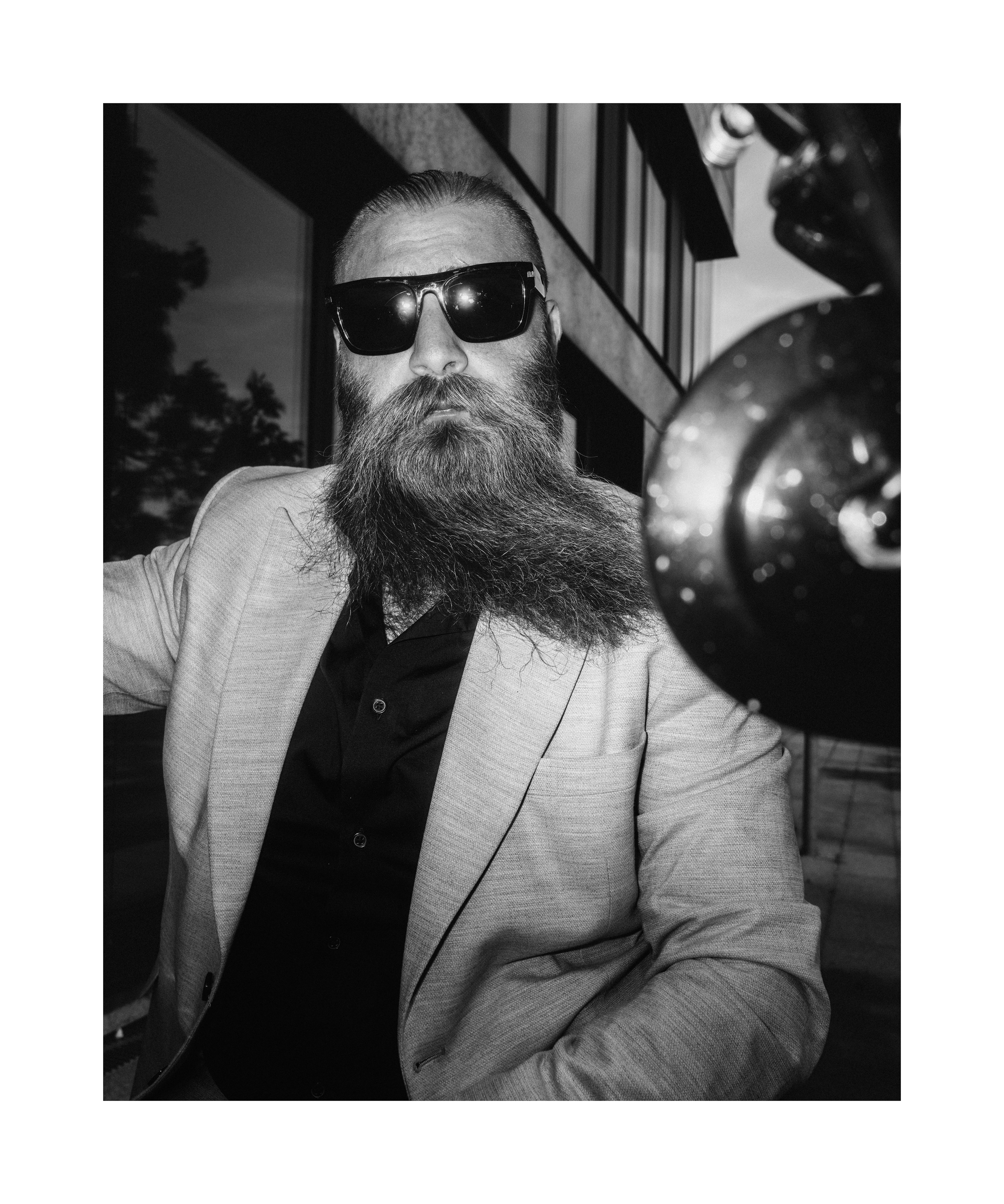 Black and white editorial portrait of a bearded man in sunglasses and a light suit beside a motorcycle.