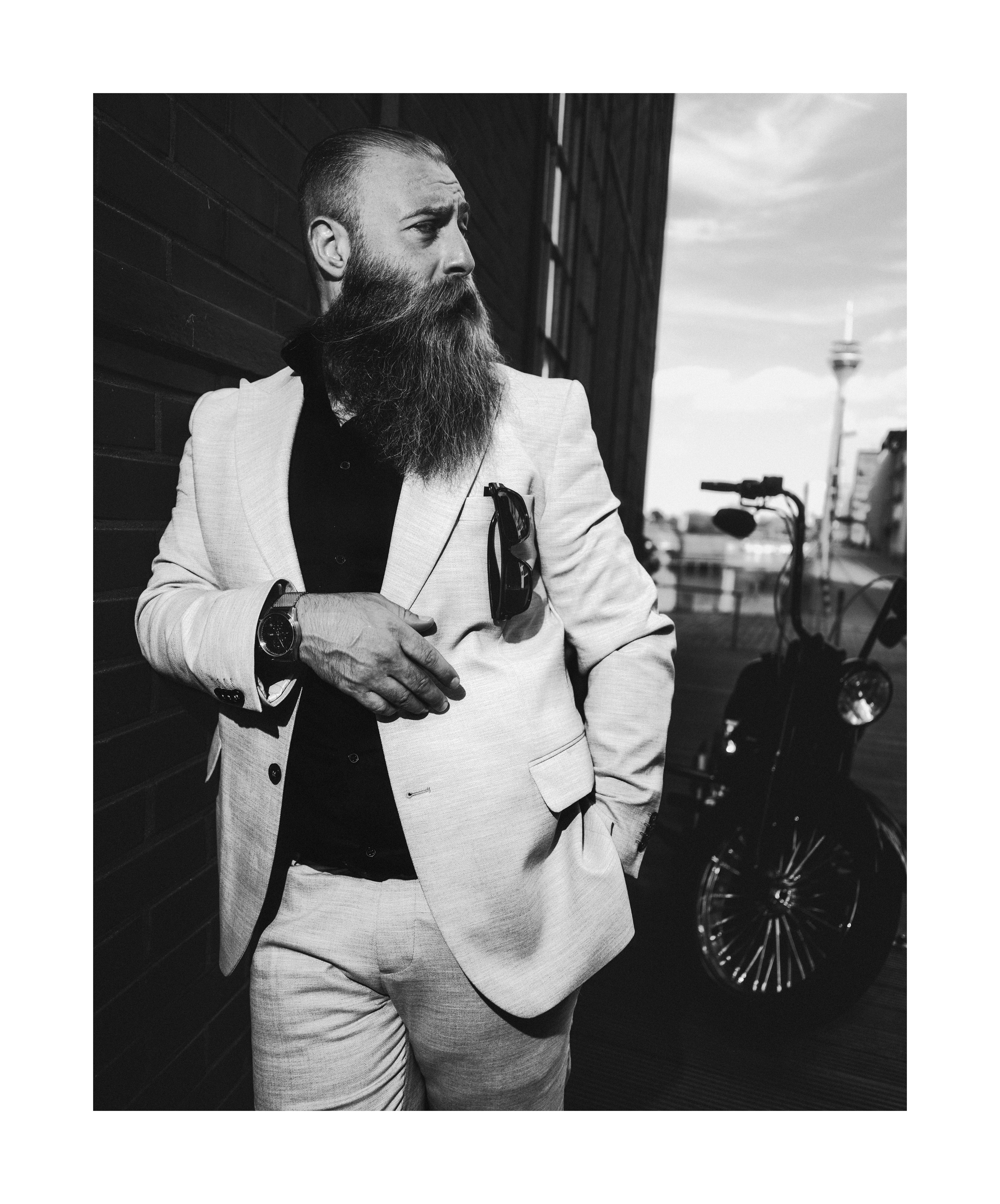 Black and white editorial portrait of a bearded man in a suit standing beside a Harley-Davidson motorcycle.