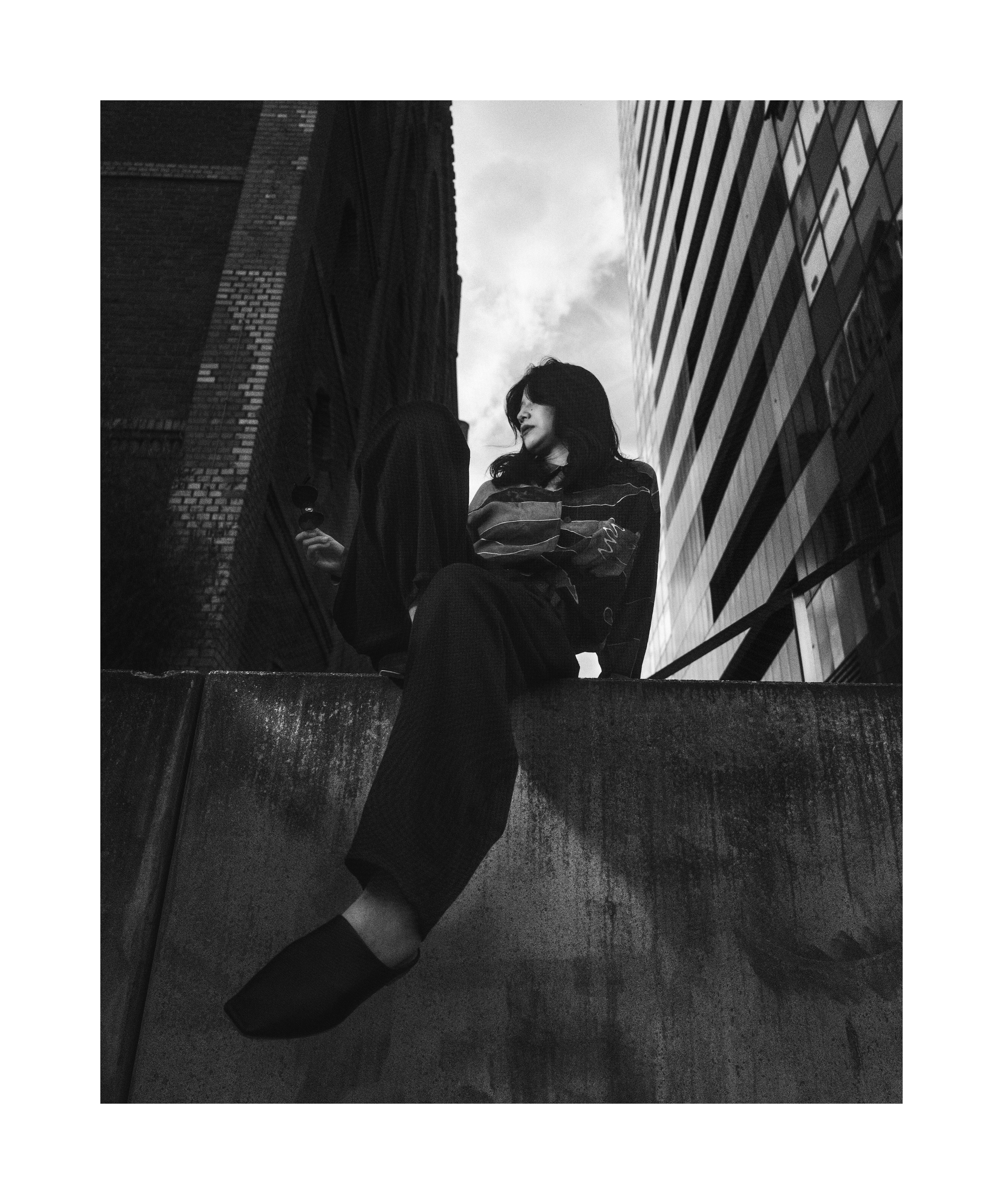 Black and white editorial portrait of Madoka sitting between urban buildings