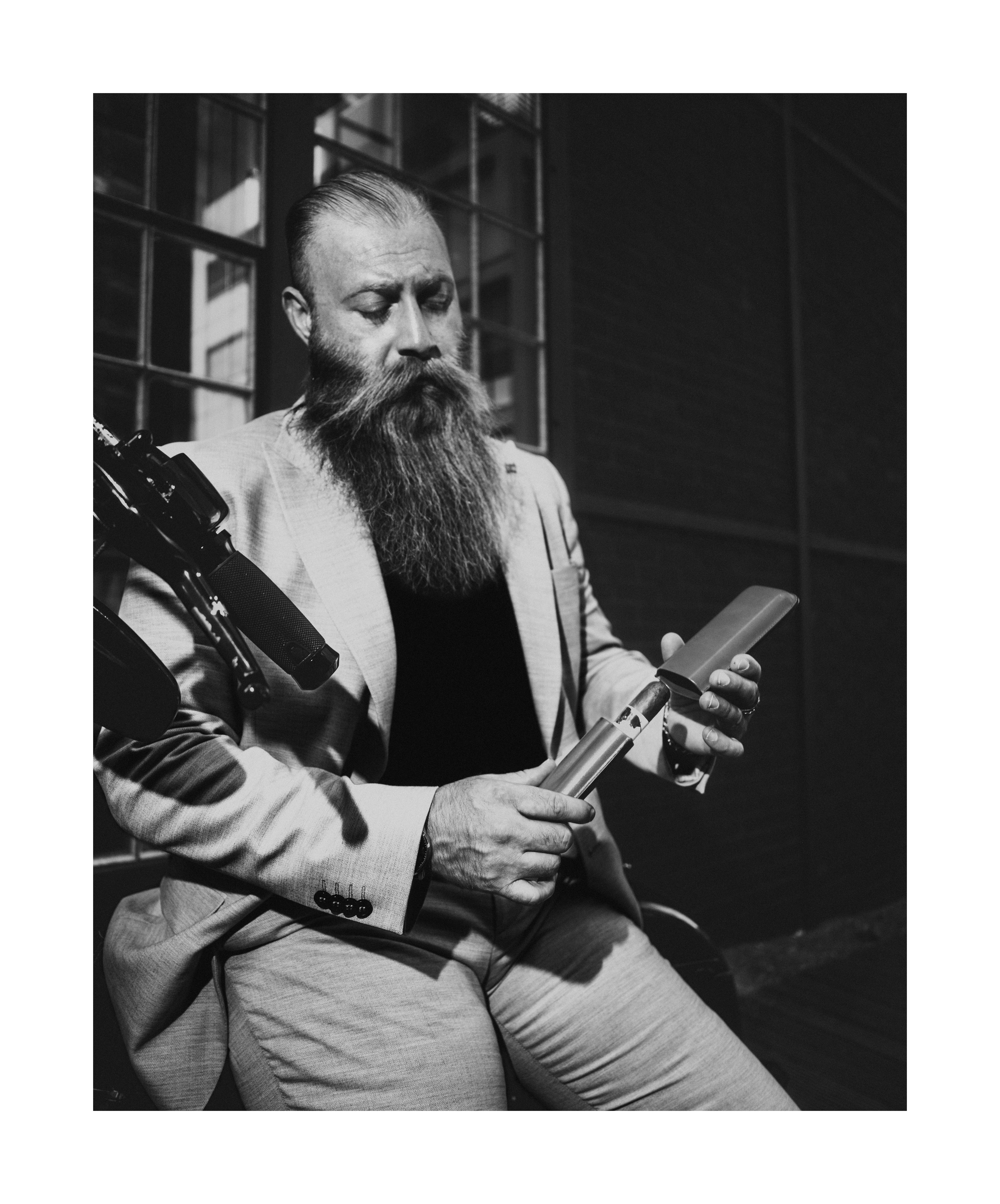 Black and white editorial portrait of a bearded man in a light suit working on a Harley-Davidson motorcycle.