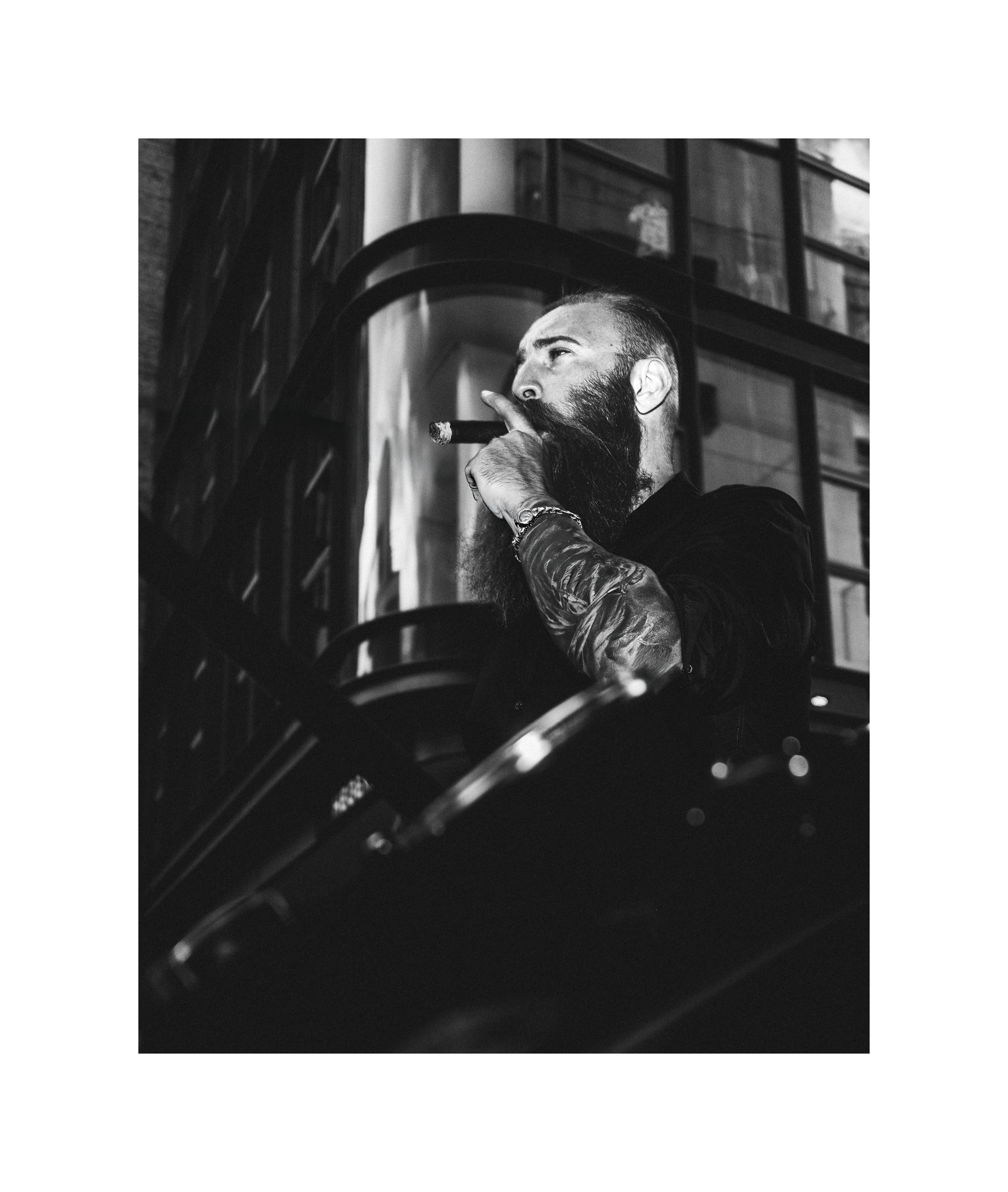 Black and white editorial portrait of a bearded man smoking a cigar beside a Harley-Davidson motorcycle.