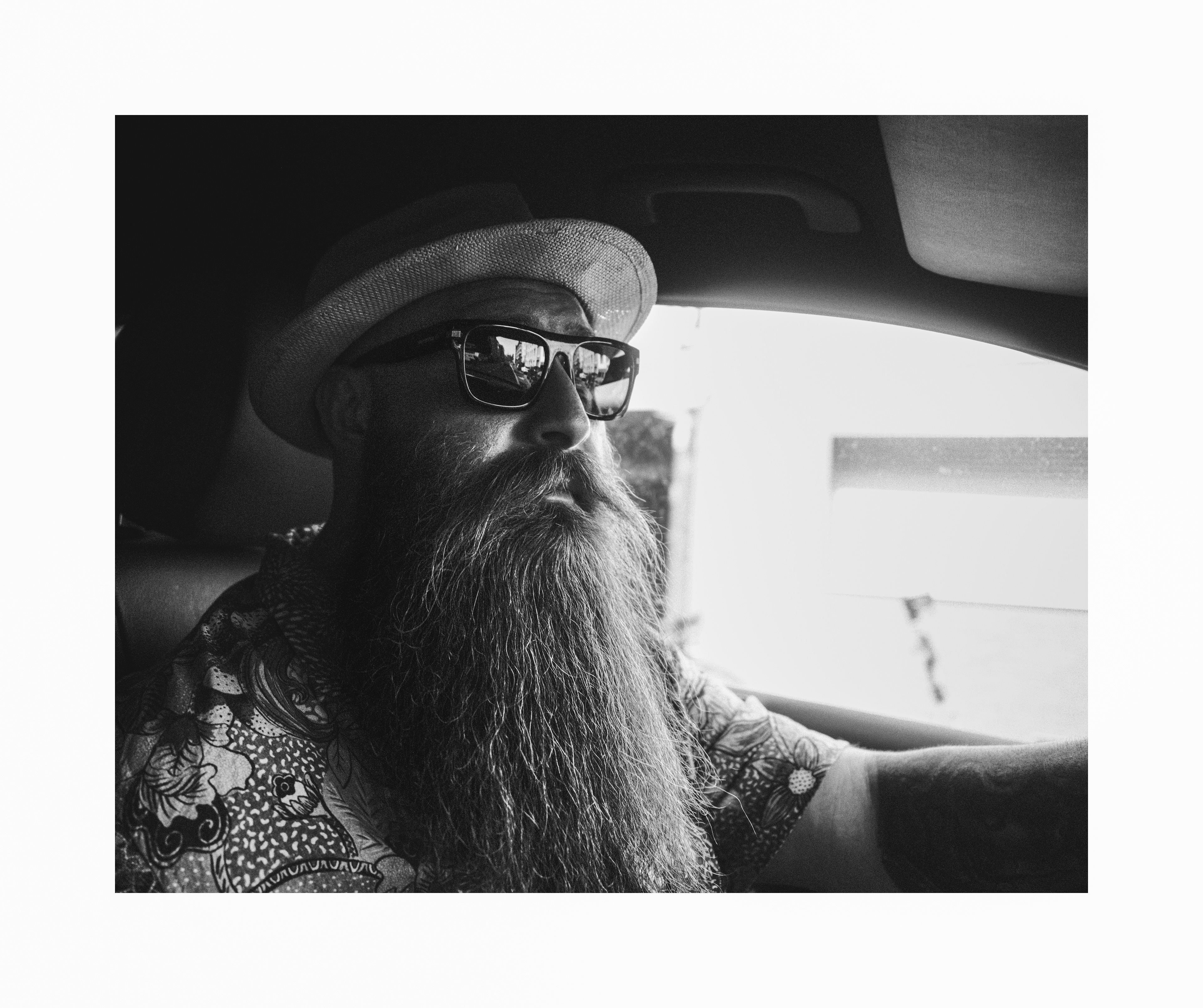 Black and white editorial portrait of a bearded man wearing a hat and sunglasses while driving