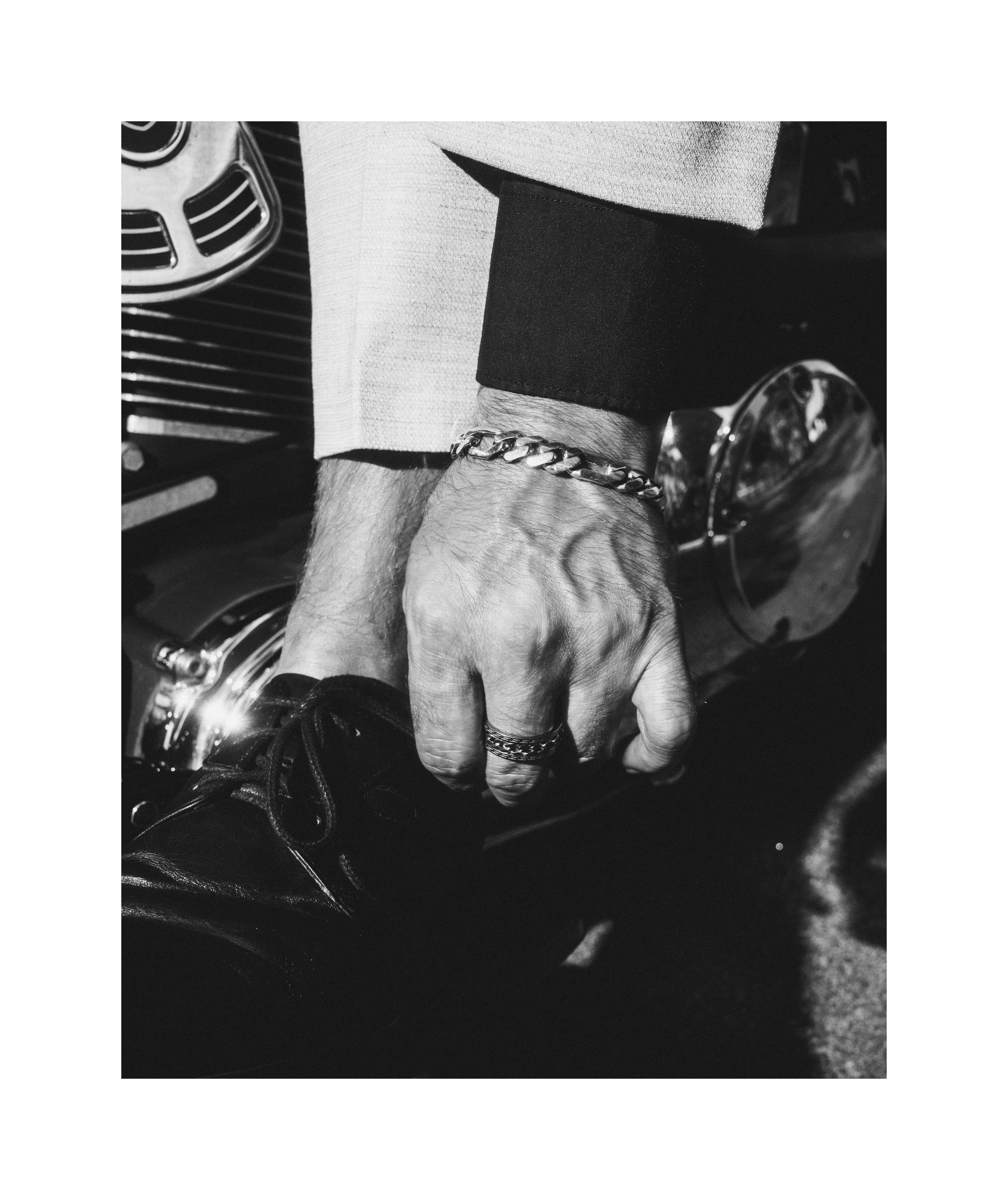 Black and white detail of a man's hand with rings and bracelet beside a Harley-Davidson motorcycle.