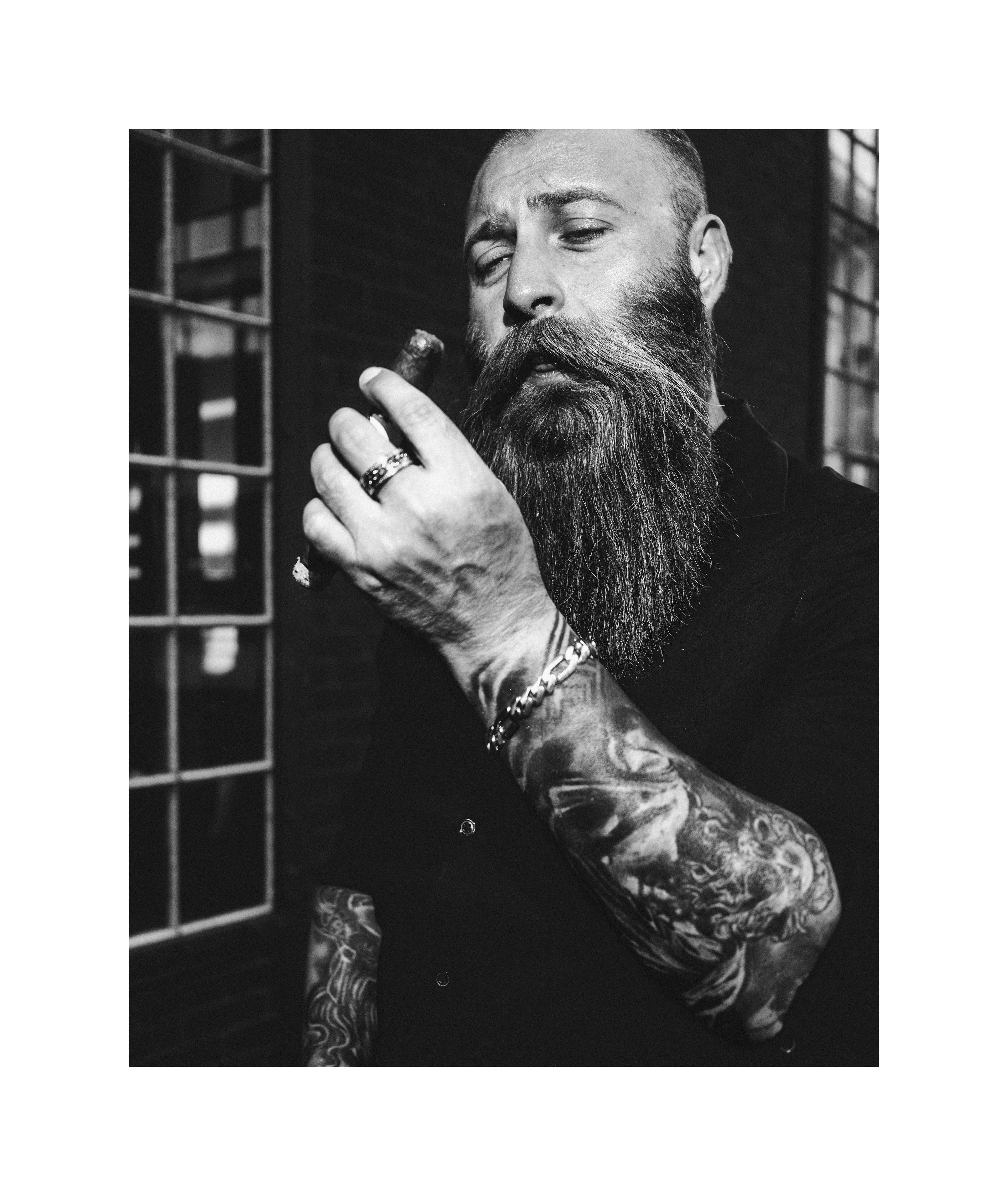 Black and white editorial portrait of a bearded man smoking a cigar, with tattoos and dramatic light.