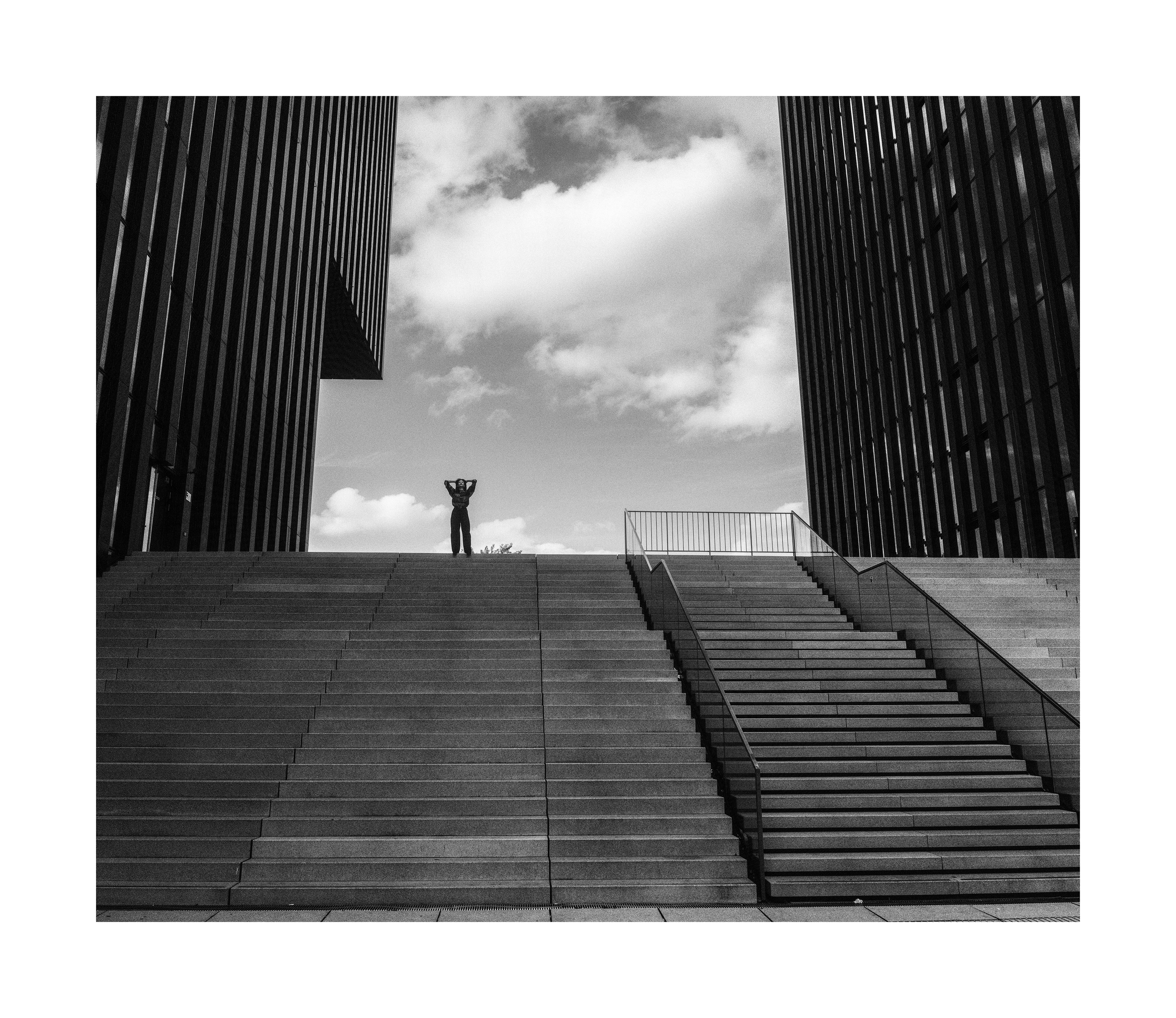 Black and white street editorial of Madoka standing on wide urban steps between modern buildings