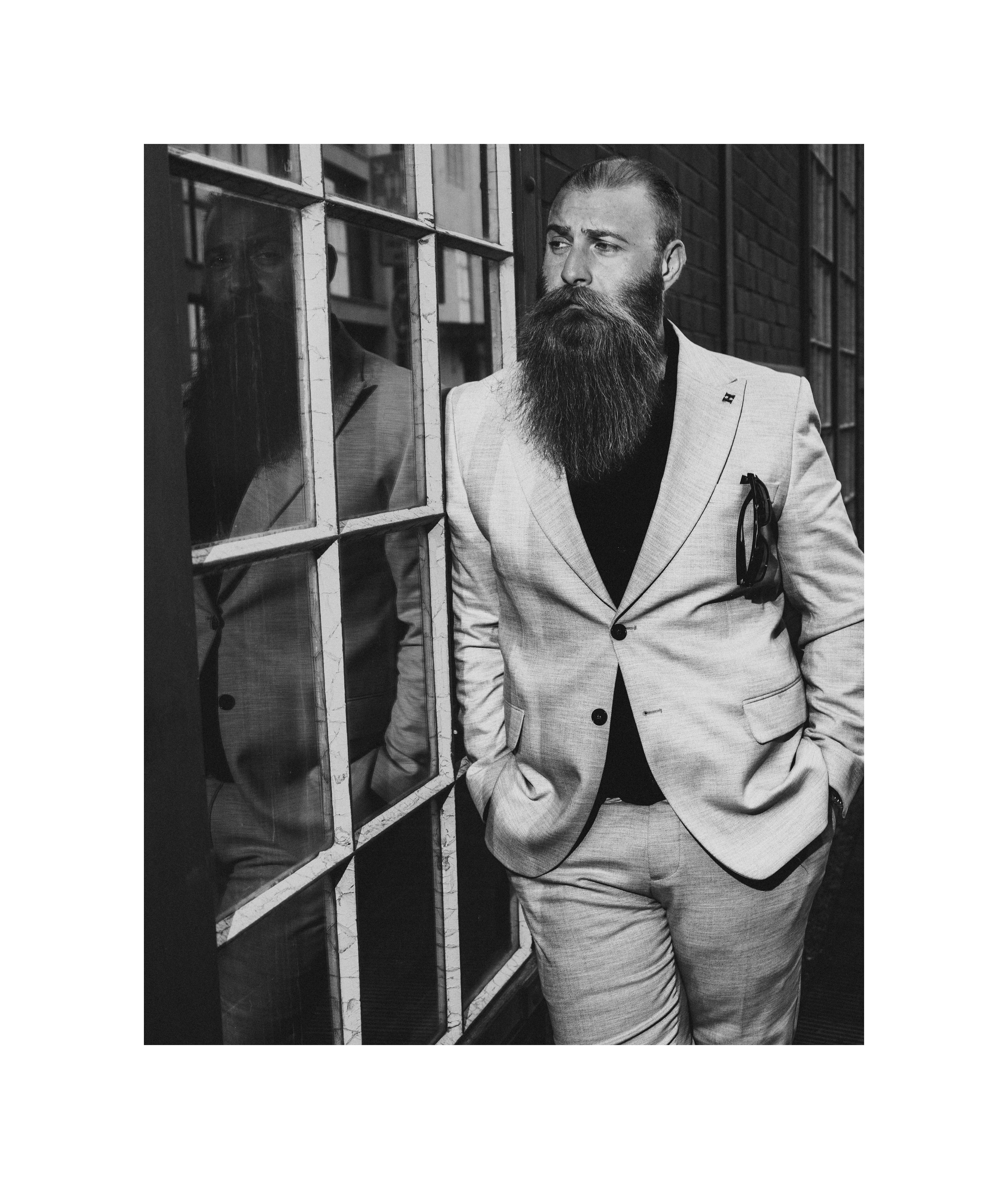 Black and white editorial portrait of a bearded man in a suit standing beside an industrial window with his reflection visible.