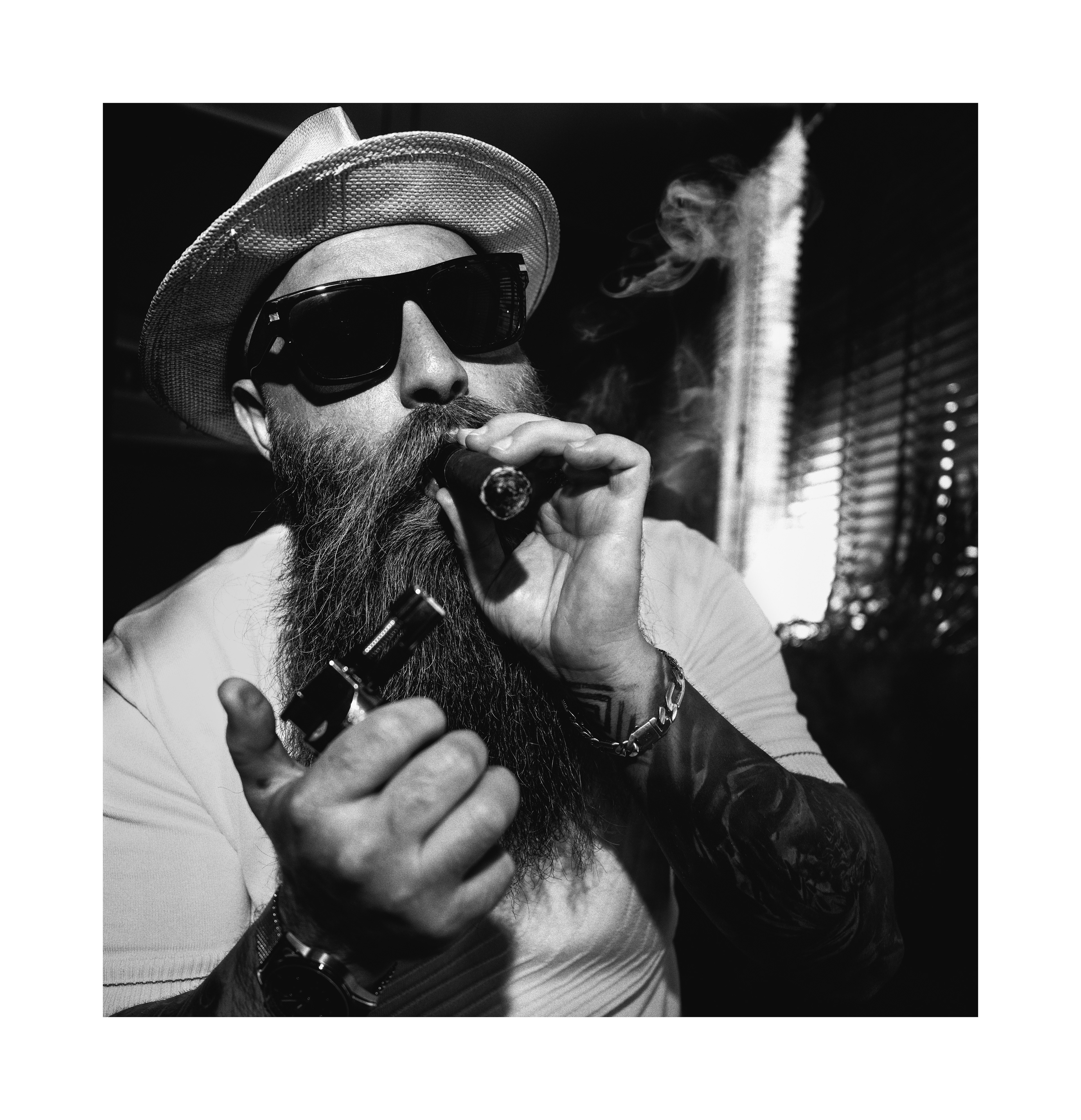 Black and white editorial portrait of a bearded man in sunglasses smoking a cigar