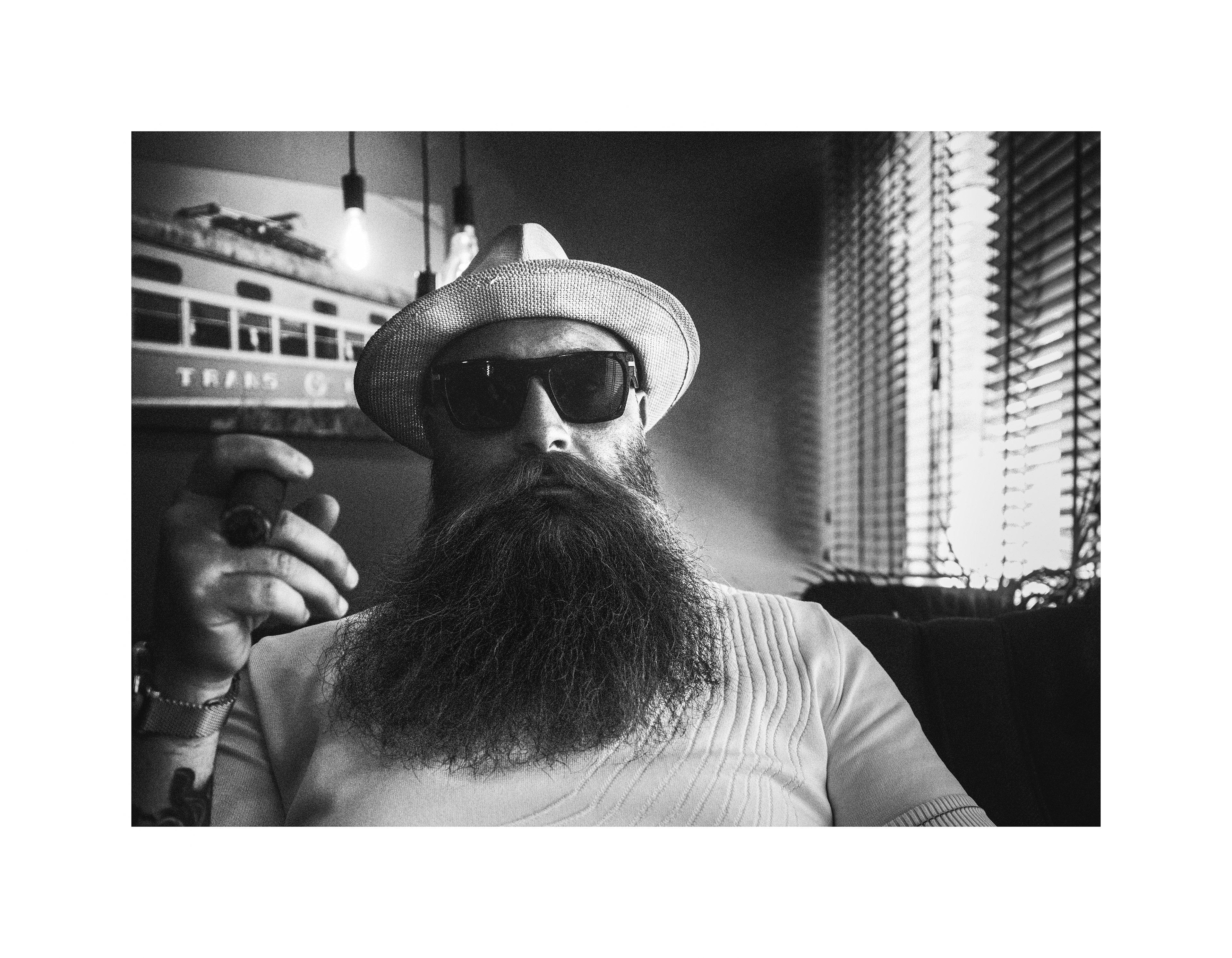 Black and white editorial portrait of a bearded man wearing a hat and sunglasses holding a cigar