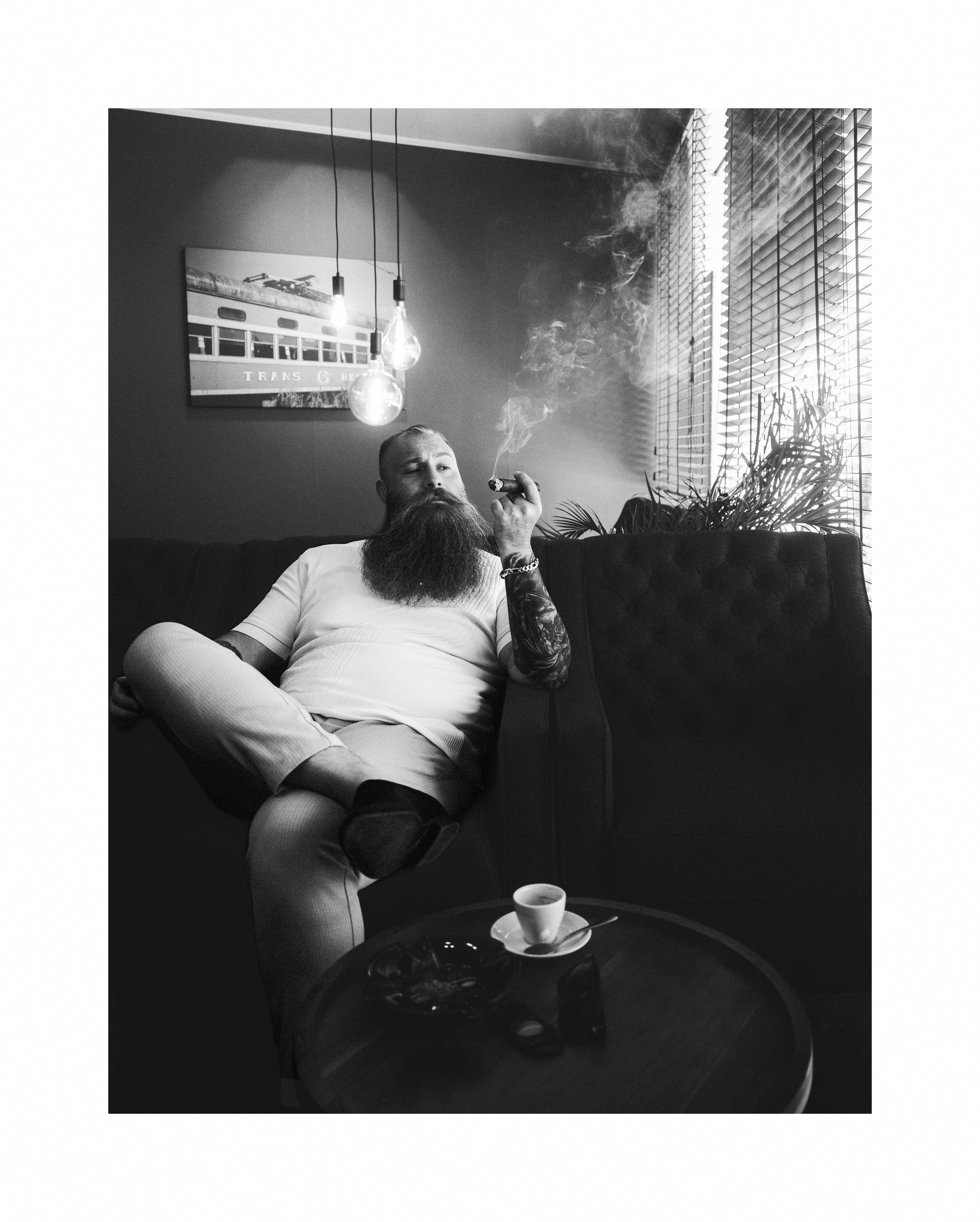Black and white editorial portrait of a bearded man relaxing in an armchair and smoking a cigar