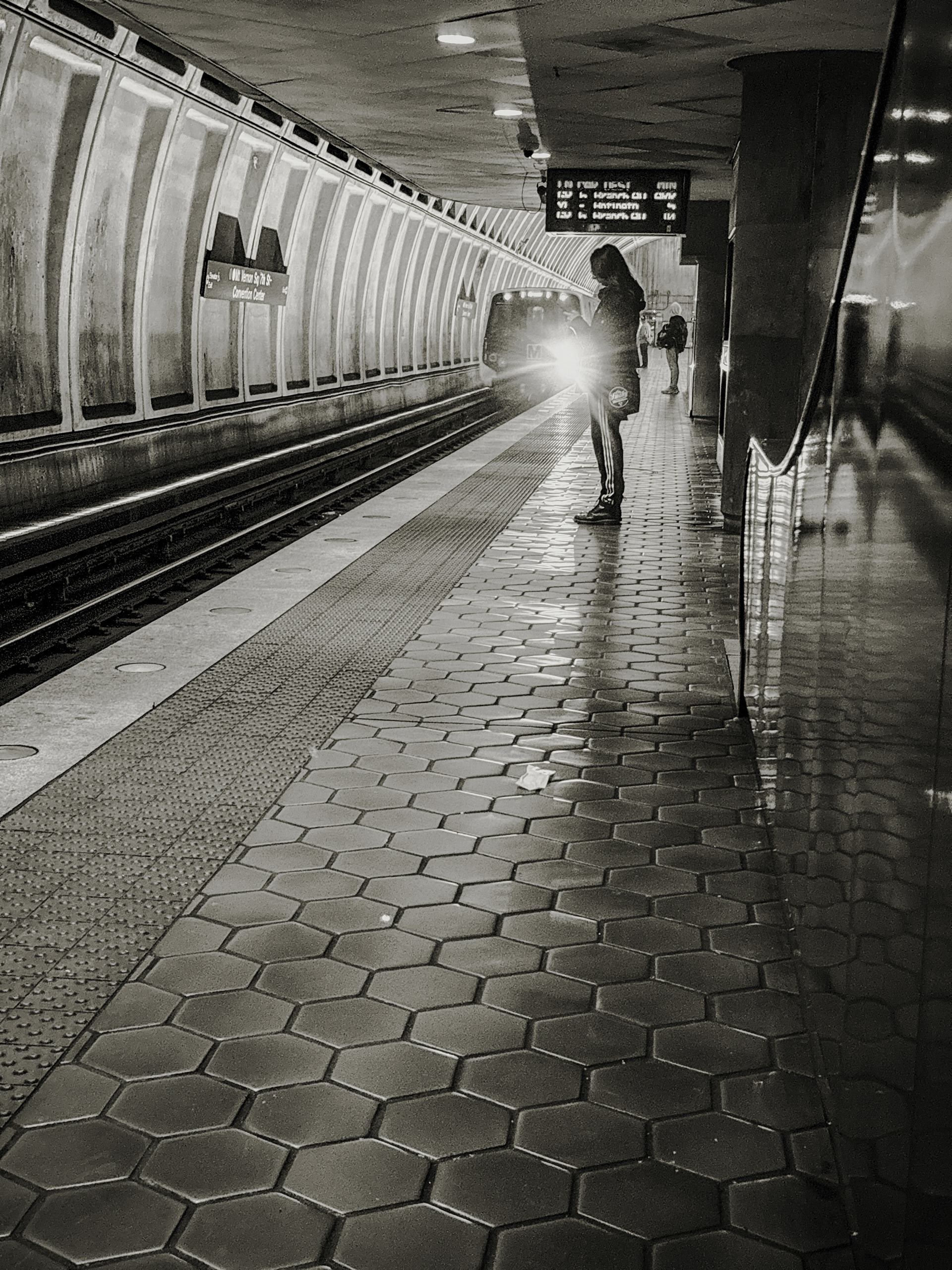 Black and white photograph of a person walking along a subway platform toward an oncoming train, its headlight illuminating the tunnel.