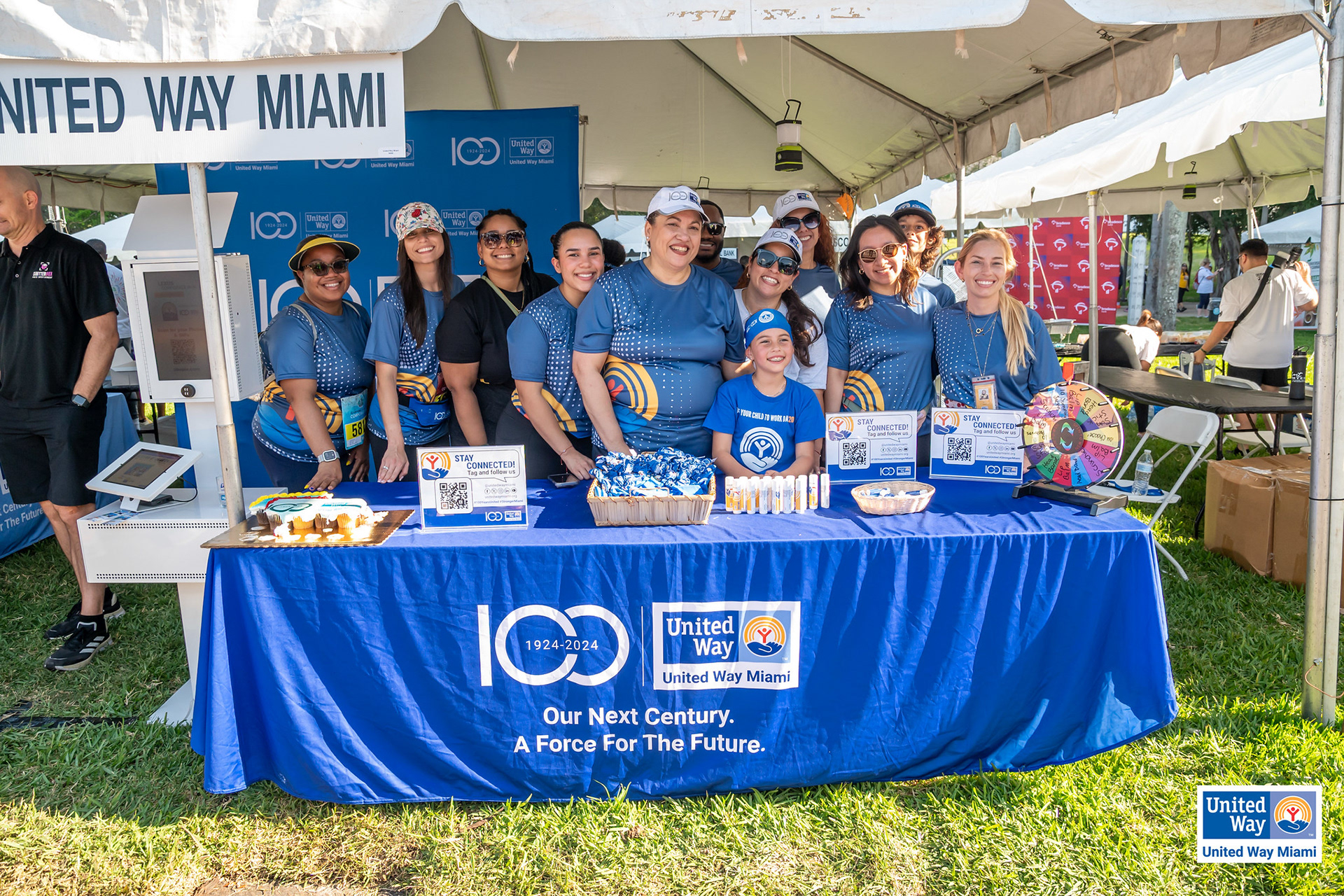 UWM 100 Years lipbalms, scrunchies and sunsticks. Staff wearing cap, headband, Dri-FIT T-Shirt and children's T-Shirt