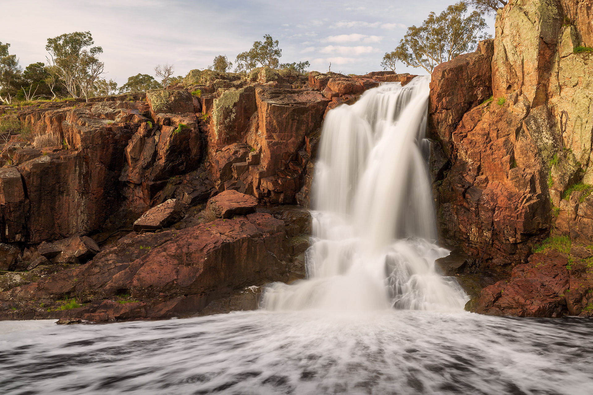 Photograph of water falling over rusty orange iron bearing rock in muted susnet light, by Dean M. Chriss..