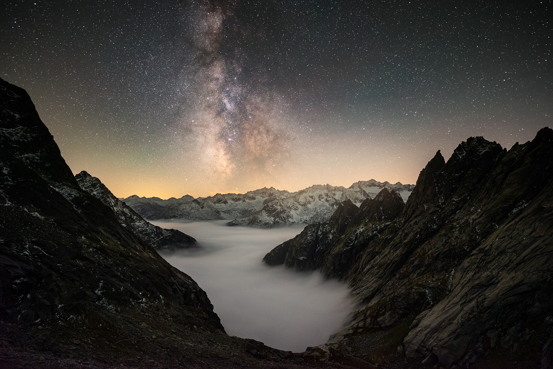 Nighttime alpine landscape with the Milky Way above a fog-filled valley surrounded by rugged mountain peaks