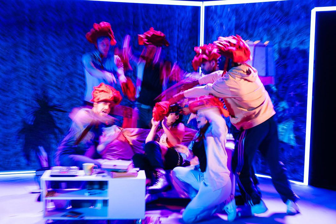 A production image from the National Youth Theatre's production of "Love Bomb" which features members of the cast with large red rose hats huddled around a character on a sofa