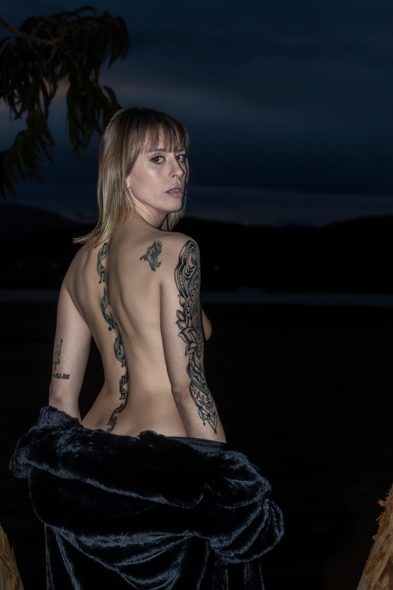Nighttime Allure: Blonde Woman in an Open Black Coat, Revealing a Tattooed Shoulder on a Beach