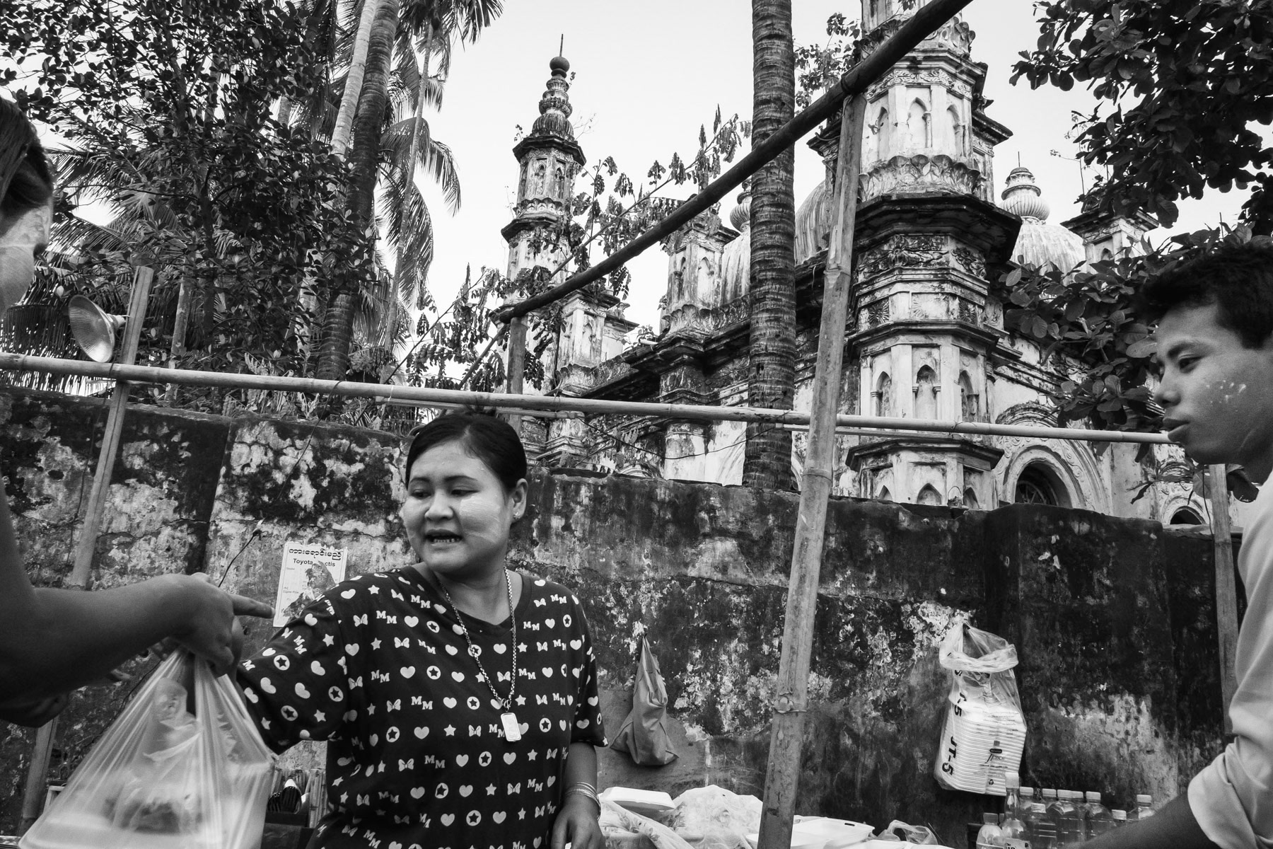 Street food stall in the capital of Rakhine, with the fenced-off mosque in the background. The mosque is inaccessible as part of a plan to erase all Muslim signs in the city. Sittwe, Myanmar, June 2016