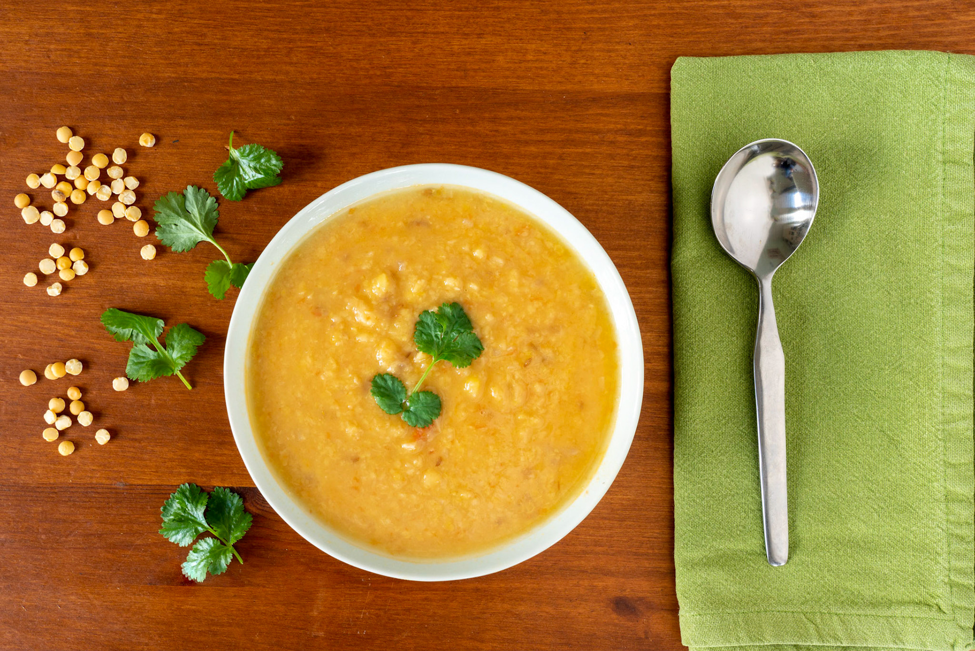 Yellow split peas lentil soup, garnished with corrinader leaves, in a bowl with a spoon and napkin on a wooden table. View from above.