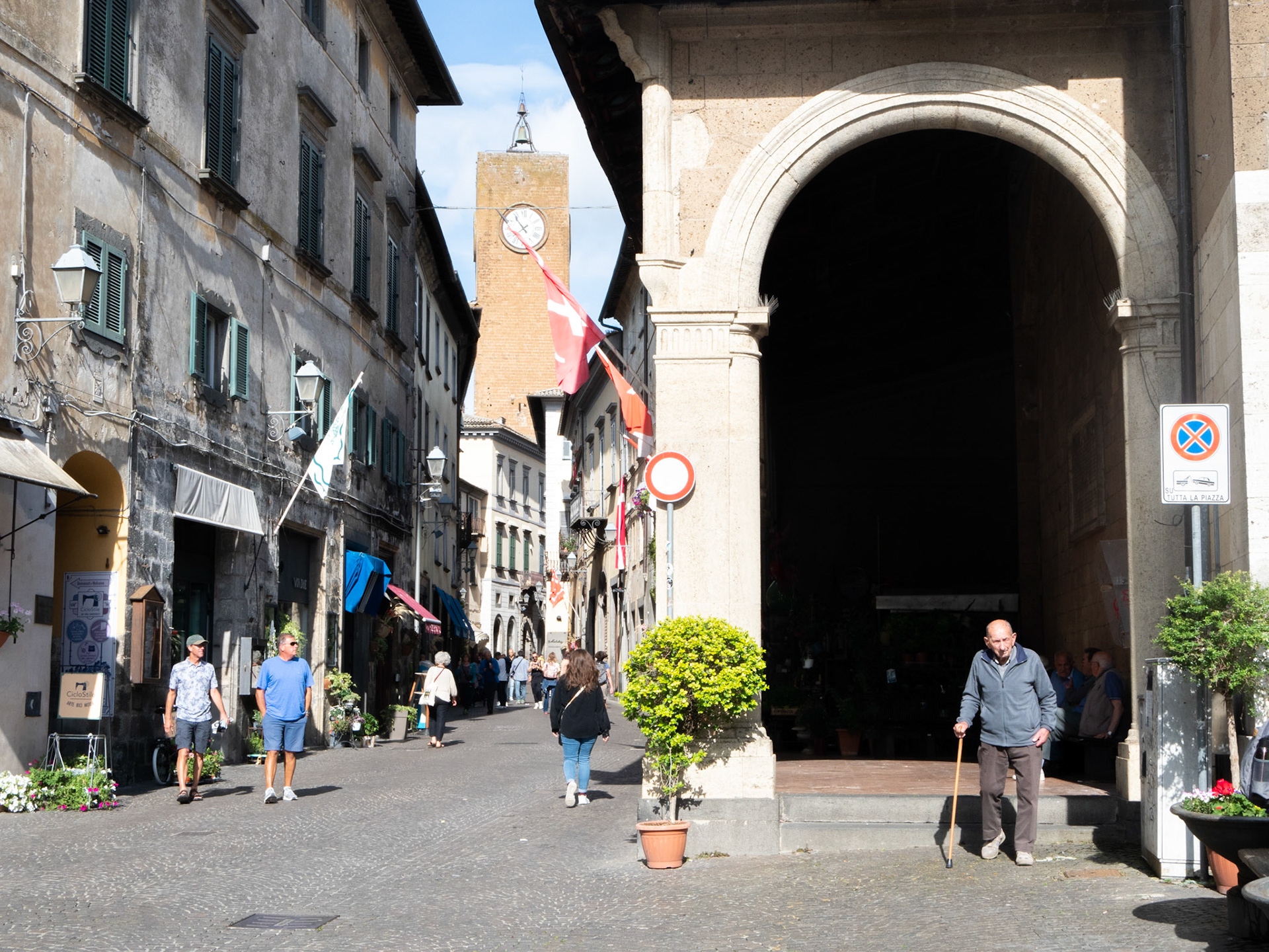 An old man walks out of an archway while a group of people talk in the background in front of the clock tower in Orvirto, Italy, on Tuesday, June 6th, 2023. Orvieto is usually busy during the middle of the day and has many tourists. (Photograph by Ben Tisdale)