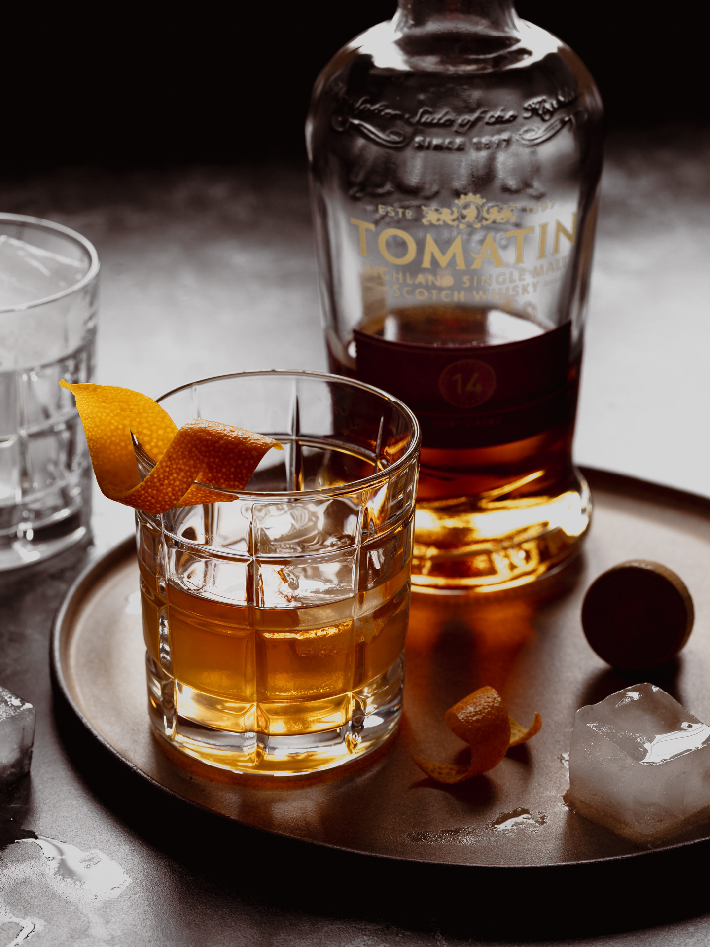 A drink photography image of whisky served in a glass with ice and garnished with orange peel made by Ioana Ursu Drink Photographer UK