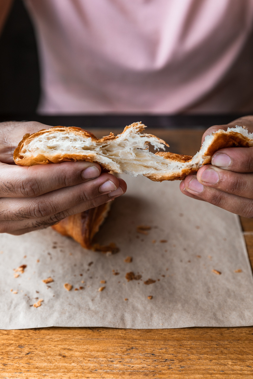 A cafe photography image made for the hospitality industry, presenting a croissant that it's being teared apart by the hands of a customer. The image is captured by Ioana Ursu restaurant and cafe photographer based in Warwickshire