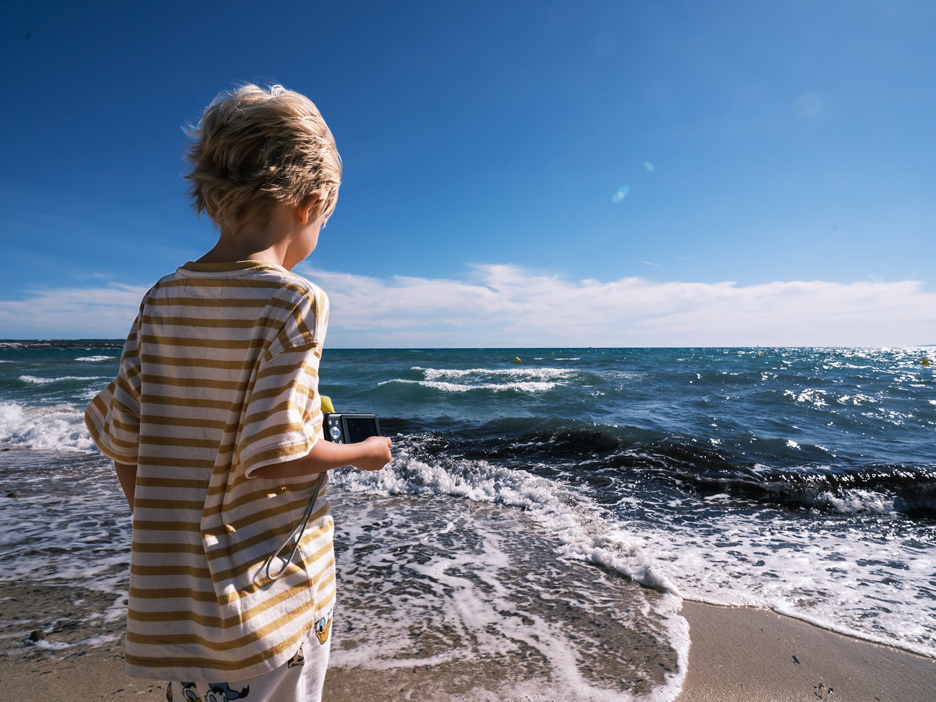 Captured on the sandy shores of Platja de Palma, this image beautifully portrays a young child lost in the wonder of the sea. Clutching a camera, the boy seems poised to capture the dynamic waves, his focus directed toward the vast, sparkling Mediterranean. His striped shirt flutters slightly in the sea breeze, blending with the blues and whites of the ocean and sky. This moment encapsulates the pure joy and curiosity of youth, exploring the natural world through his lens. It's a vivid reminder of the early adventures that shape our understanding and appreciation of the environment around us.