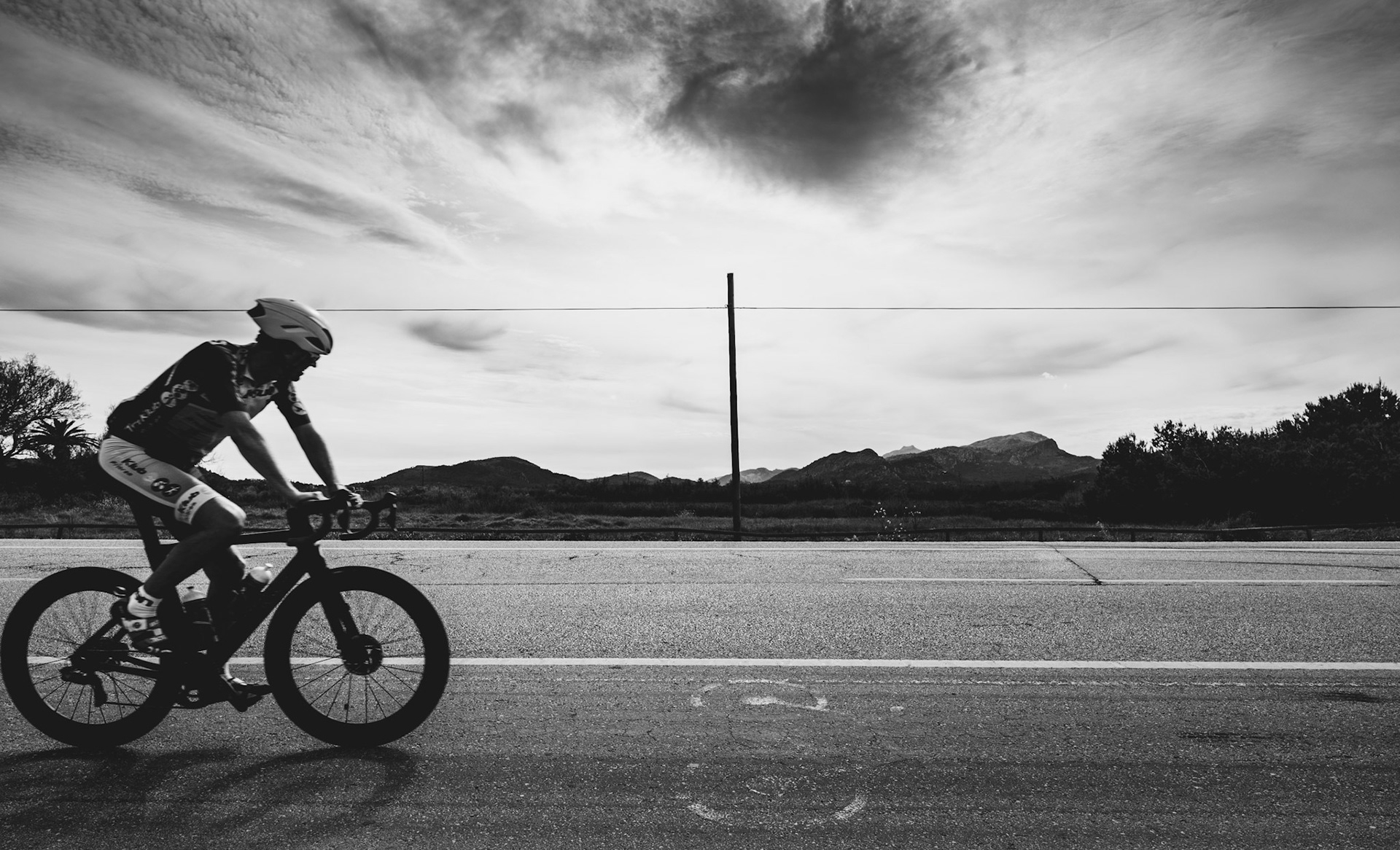 this photograph captures a cyclist in mid-ride on the road that connects pollenca with alcudia in mallorca. the cyclist, dressed in full gear and focused on the journey, is framed against a dramatic sky. the image, taken in black and white, emphasizes the textures of the road and the cloud formations above. the road itself is known for its scenic views, with the sea just behind the photographer, and the landscape offering a mix of coastal beauty and rural tranquility.