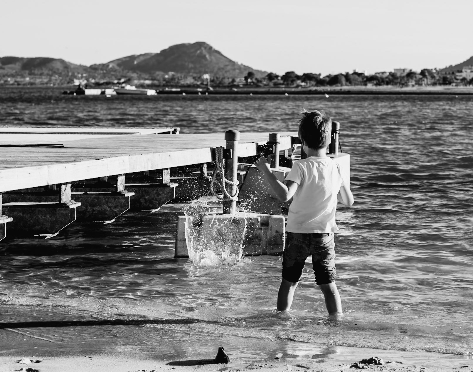 in the harbor of pollença, a young boy becomes the focal point of a scene washed in the crisp light of innocence. the black and white tones strip away the distractions, leaving us with the universal language of childhood—a moment of pure joy, hands casting sand into the sea. the mountains and boats in the distance stand as silent witnesses to this simple act, timeless and free. it’s a dance with the elements, with each grain of sand a note in a symphony of playfulness, and the water, an eager partner, catches the offerings in a splash of sparkling laughter.
