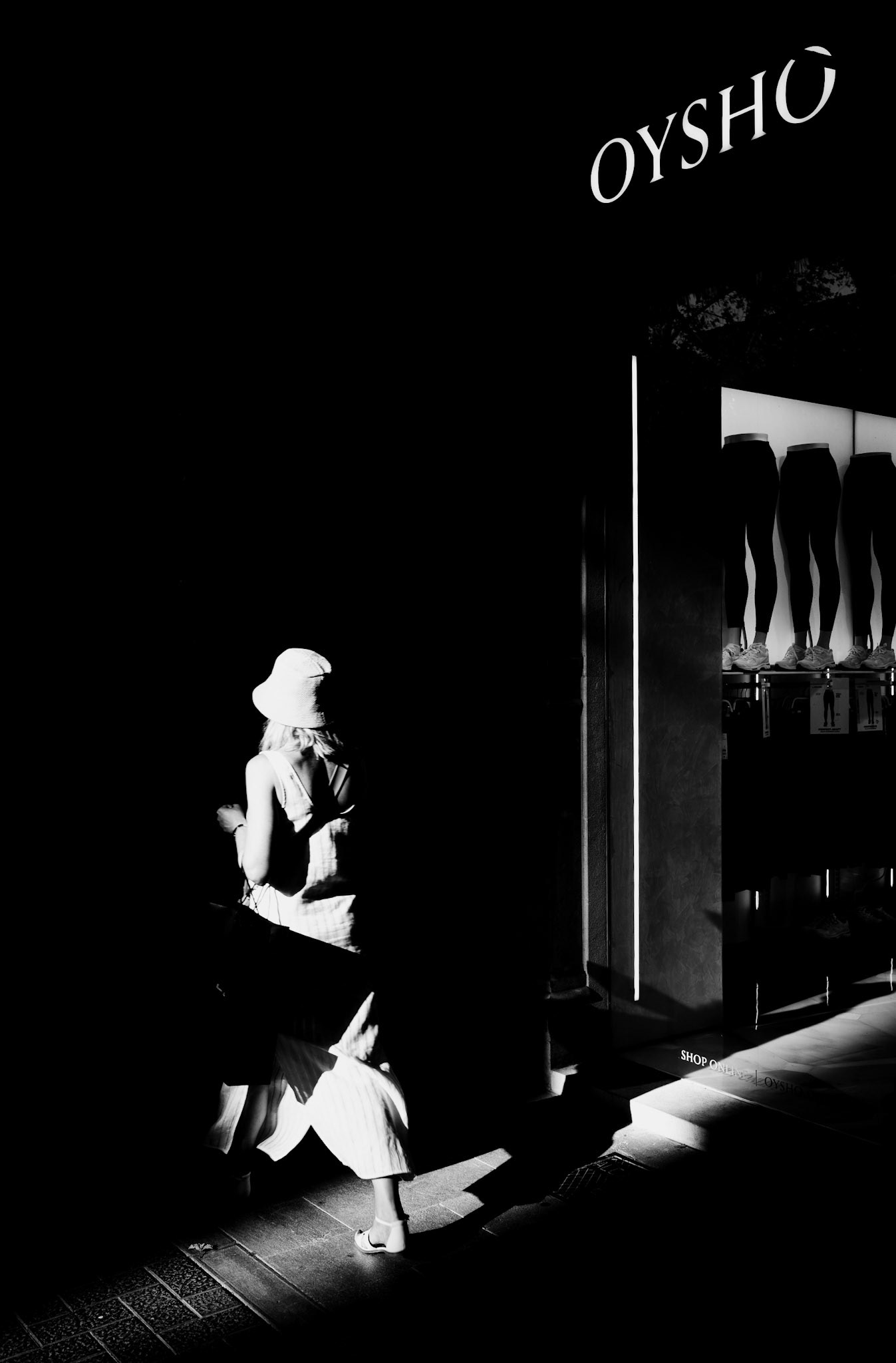As the evening sun dipped low on Palma's Paseo del Borne, I captured this fleeting moment of a woman seemingly lost in her thoughts, stepping through the interplay of light and shadow. The golden hues of the setting sun illuminate her white dress, casting a serene glow that contrasts sharply with the darkness surrounding her. The minimalistic storefront of Oysho in the background adds a modern touch, while the shadows almost create a frame, isolating her in a moment of quiet elegance. It’s a snapshot of the everyday, turned almost cinematic by the magic of the golden hour.