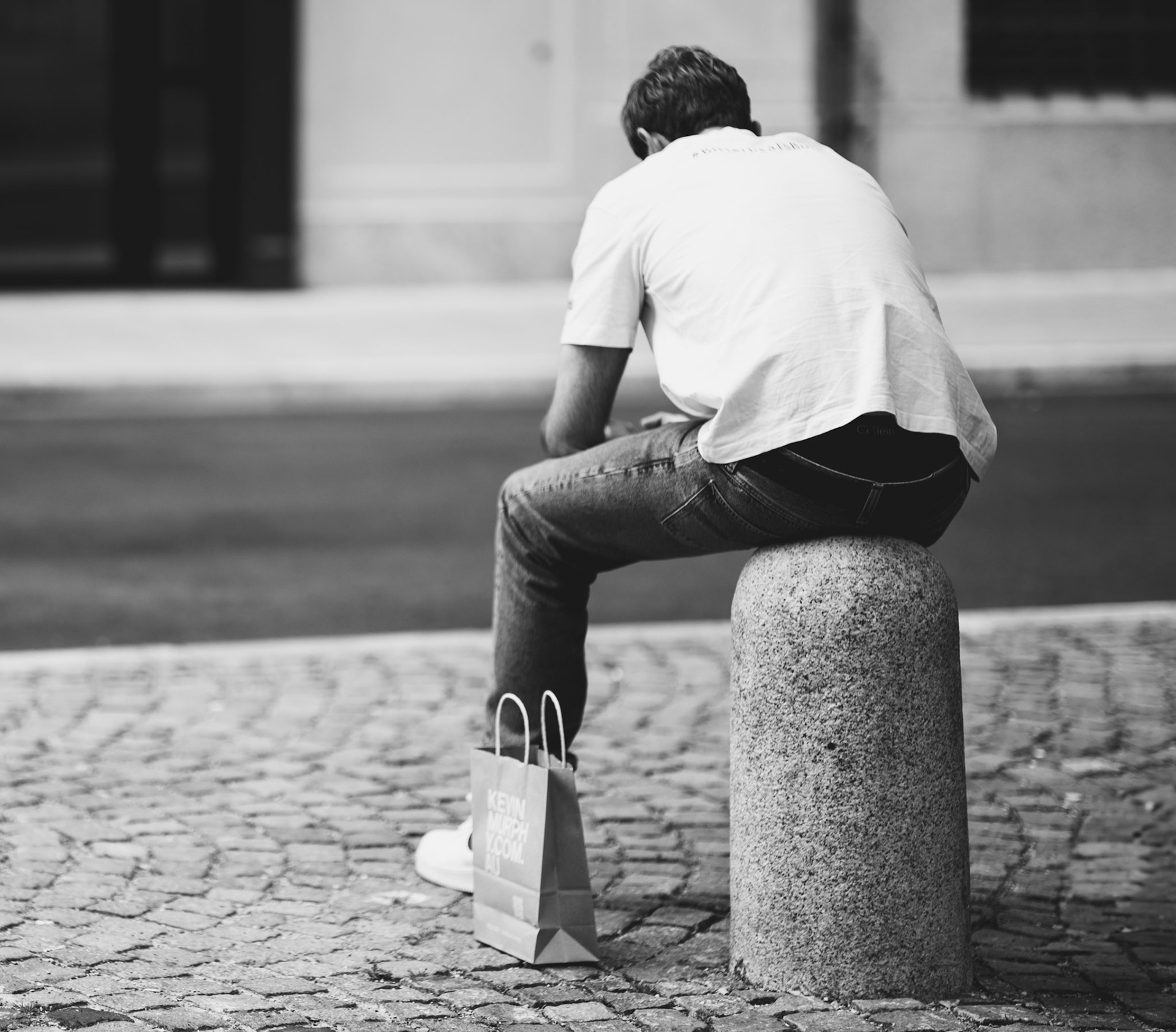 captured during a photowalk in munich, this image encapsulates a quiet moment of rest and introspection. a man sits on a stone bollard, using it as a makeshift seat, holding a shopping bag with the words "kein mensch ist illegal" (no human is illegal). the black and white photo enhances the contours and textures, with the cobblestone street and the unassuming backdrop of urban everyday life. the man's posture and the stillness of the street reflect a snapshot of modern city life, where every moment can be a pause for contemplation and reflection.captured during a photowalk in munich, this image encapsulates a quiet moment of rest and introspection. a man sits on a stone bollard, using it as a makeshift seat, holding a shopping bag with the words "kein mensch ist illegal" (no human is illegal). the black and white photo enhances the contours and textures, with the cobblestone street and the unassuming backdrop of urban everyday life. the man's posture and the stillness of the street reflect a snapshot of modern city life, where every moment can be a pause for contemplation and reflection.