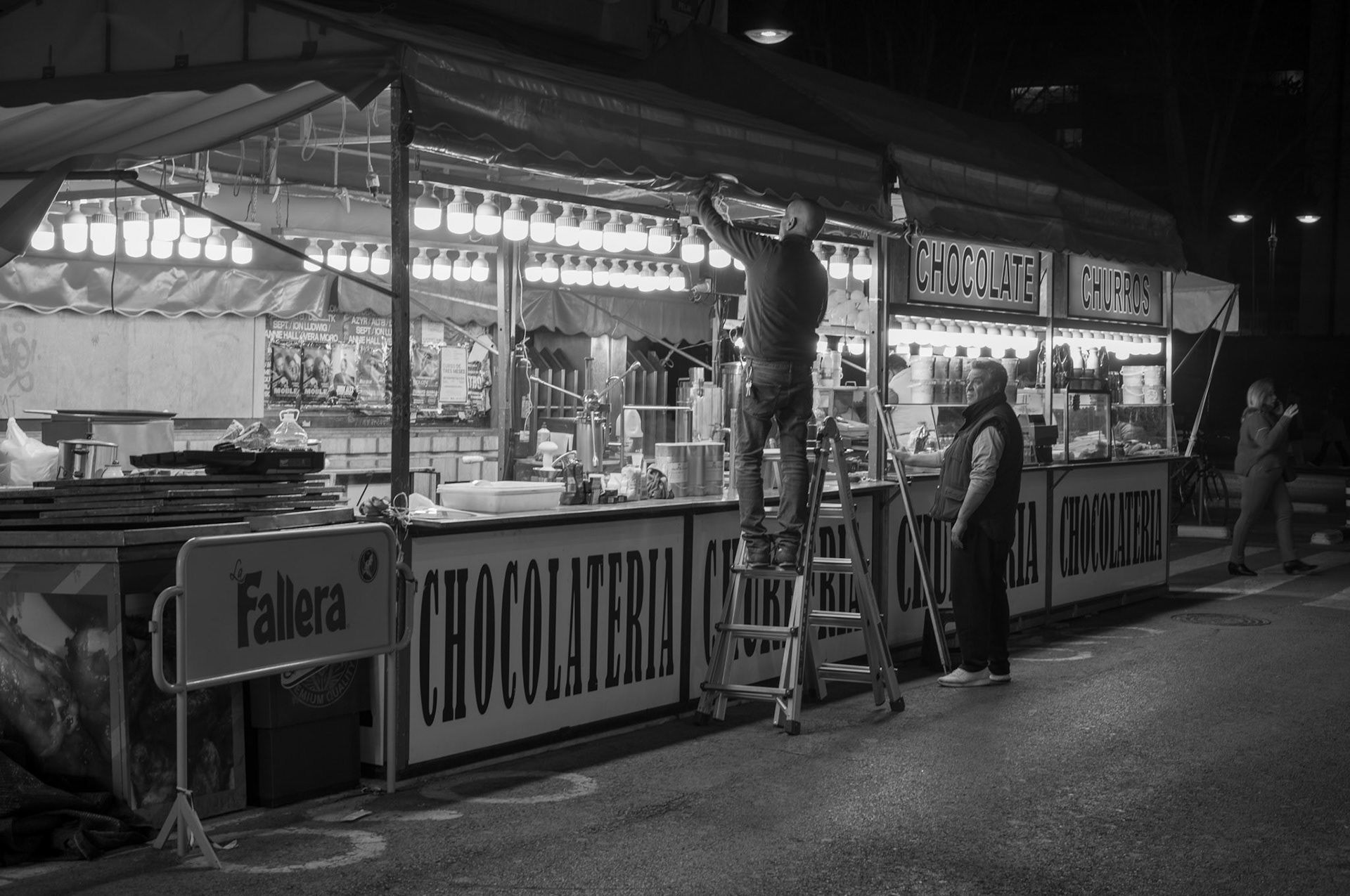 in the embrace of early nighttime in valencia, the churrería stands silent before the impending symphony of chatter and sizzling oil. a man perches atop a ladder, like a conductor poised before an orchestra, his silhouette etched against the soft glow of stark white bulbs. below, his partner watches, grounded and vigilant, a silent guardian of the soon-to-be bustling stall. the scent of chocolate and dough seems to linger in the air, awaiting the warmth of the morning rush, the clink of cups and the laughter of the day to come. this stillness speaks of anticipation, of moments just before the curtain rises, where every light and shadow holds the breath of stories yet to unfold.