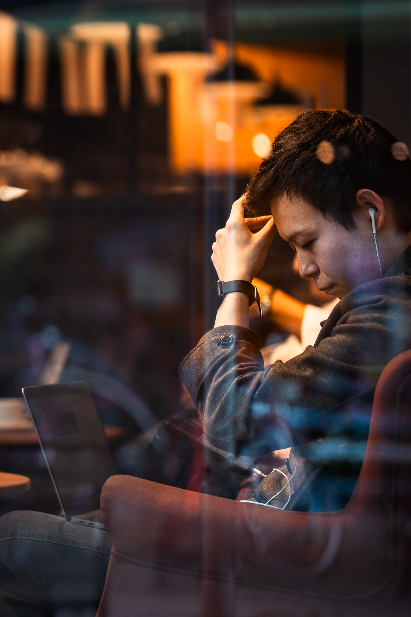 captured through the café window in berlin mitte, this image reveals a young man immersed in his own thoughts, separated from the bustling world outside. the warm reflections and soft colors of the café atmosphere evoke a sense of calm and introspection, as if he’s momentarily detached from his surroundings. the glass creates a quiet barrier between observer and observed, between outside and inside, while the light gently highlights the contours of his face and hand. this moment of silent reflection tells a small, personal story amidst the big city, as if the world around him has paused for just a second.