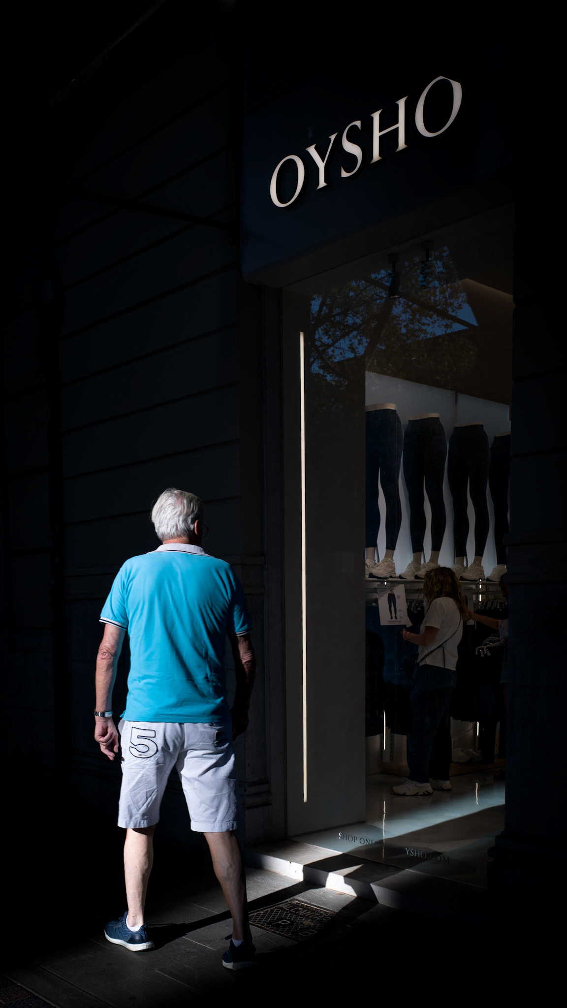 In the early evening light on Palma's Paseo del Borne, the scene captures an intriguing moment of indecision. A man, drawn to the allure of the Oysho store, stands transfixed but does not venture inside. The contrasting interplay of light and shadow adds to the mysterious atmosphere, as if the man is caught in a fleeting moment of contemplation. His bright blue shirt contrasts sharply with the store's modern façade, creating a visual tension between his presence and the commercial world that beckons him.