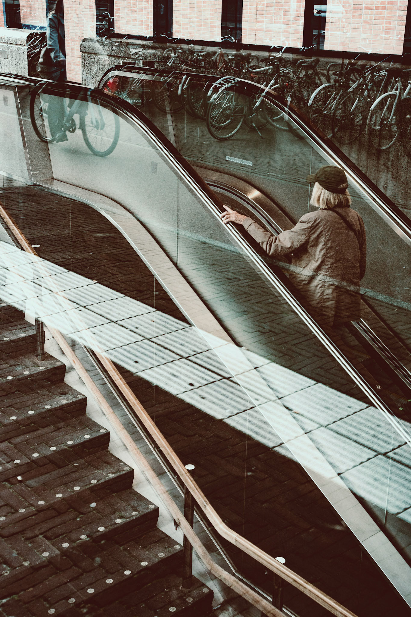 amidst the quiet motion of the city, a lone figure ascends on the escalator, their reflection melding with the bicycles parked above, creating layers of daily life. the soft shadows and lines crossing the glass give the image a dreamy, introspective tone. a pause in the rhythm of the day, where the reflections mirror a moment of stillness amidst the flow of urban life.