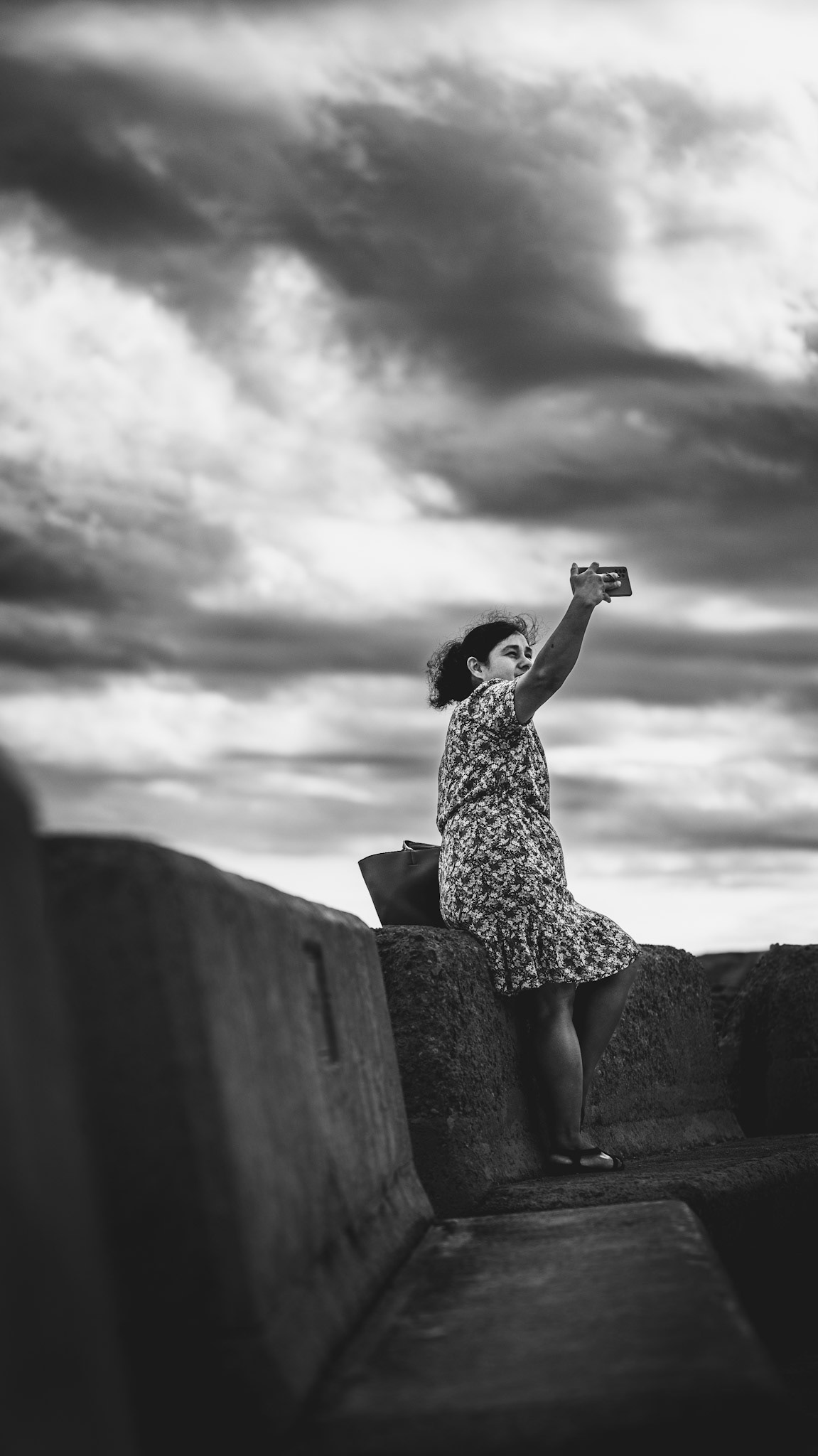 captured on a windy autumn day in portixol, this black-and-white shot shows a woman raising her phone towards the cloudy sky. sitting on a weathered stone wall, she looks focused, perhaps trying to capture the moment despite the stormy weather. the dramatic sky and the strong contrast between her floral dress and the heavy clouds create a striking, minimalist composition that highlights the raw coastal atmosphere of this breezy day.