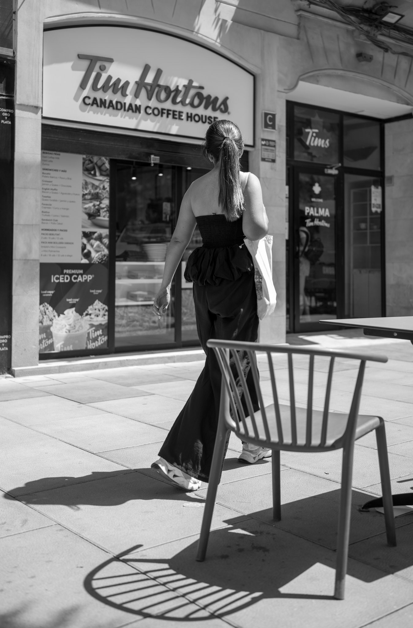 captured at plaça espanya, this image shows a woman walking past a tim hortons, seemingly contemplating whether to stop for a coffee. her stride suggests a moment of decision, ultimately choosing to continue on her way, leaving the chair empty. the shadows cast by the chair and the woman add depth to the scene, highlighting the transient nature of choices and the quiet moments in a bustling city.