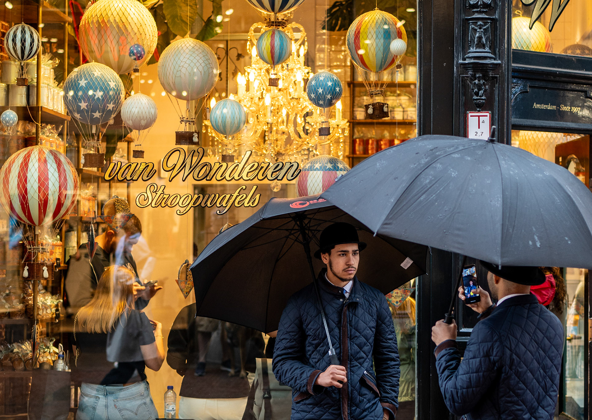 captured in the heart of amsterdam, this intriguing scene shows two men, almost mirror images of each other, standing outside a stroopwafel shop. dressed in identical quilted jackets, bowler hats, and each holding a black umbrella, one man photographs the other, creating a fascinating interplay between the photographer and the subject. the shop's whimsical hot air balloon decorations provide a vibrant backdrop, adding an unexpected twist to this candid street portrait. the image plays with themes of symmetry, identity, and the blending of traditional and modern styles in urban life.