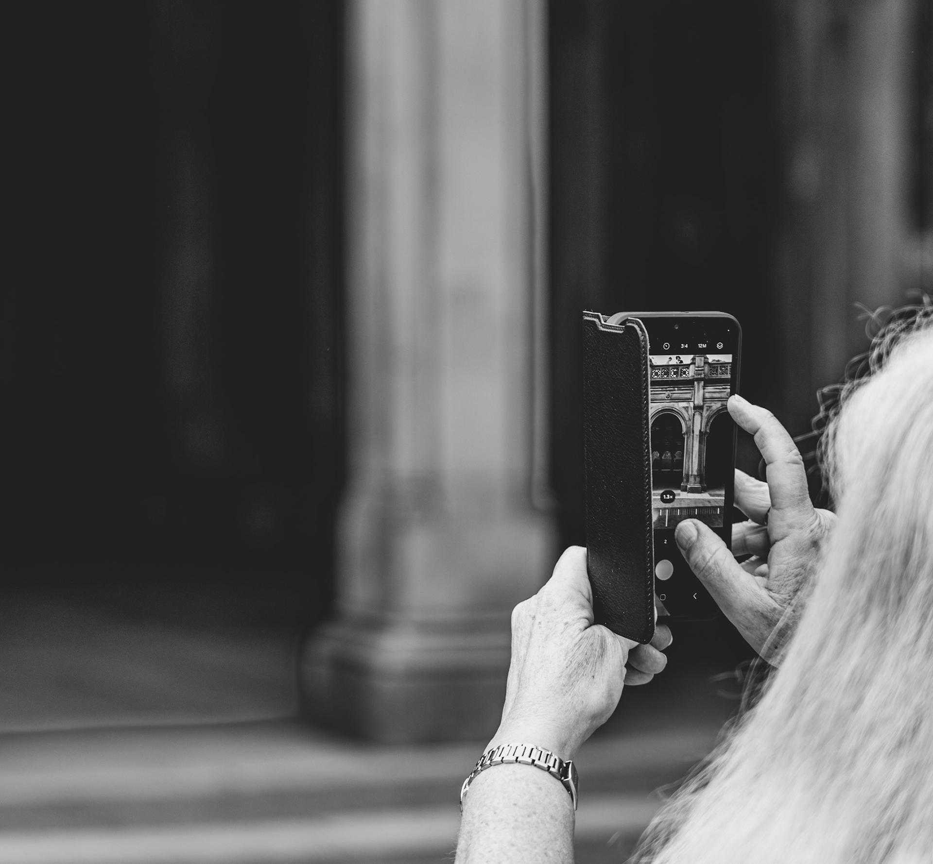 captured at bethesda terrace and arcade in central park, this photograph uniquely highlights the beautiful architecture through the lens of an iPhone held by an observer. the intricate details of the historic arches are mirrored in the modern device, creating a compelling contrast between old and new. the black and white treatment further emphasizes the timelessness of the scene, focusing on the textures and lines that define this iconic new york city landmark.