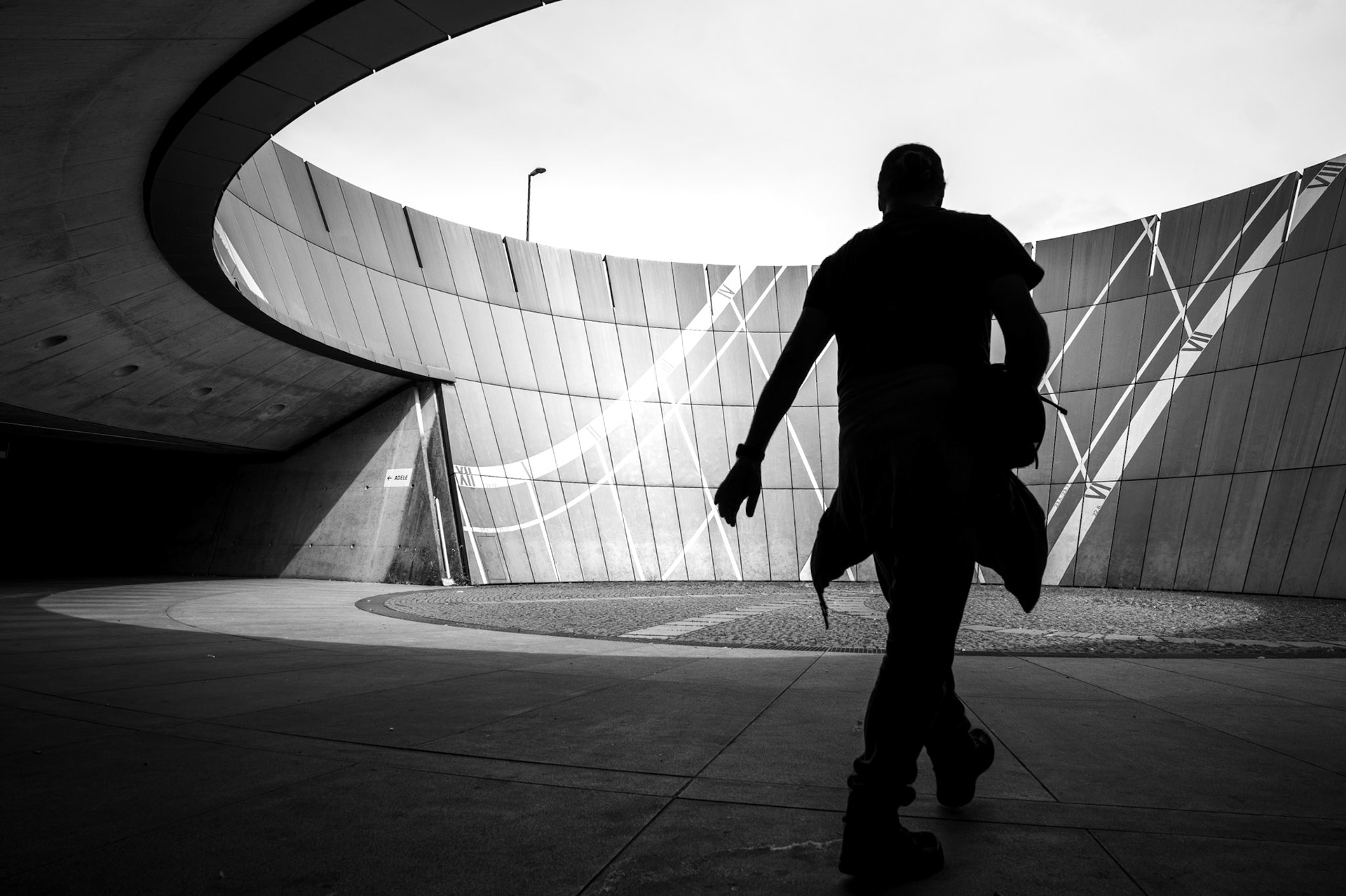 at the kunst rondell near the messestadt ost station, architecture and motion collide in a quiet choreography. the massive curve of the structure wraps around the figure, a silhouette in mid-step, frozen in a moment of quiet determination. light cuts across the metallic facade, tracing sharp, geometric lines that contrast the softness of the figure’s movement.the space feels monumental yet human – a balance between the overwhelming scale of the architecture and the fleeting presence of a person walking through it. the shadows stretch long, the curves cradle the light, and the figure disappears into the moment, leaving only the interplay of shapes and reflections behind.this is more than a photograph. it’s a scene where stillness and motion coexist, where concrete and light tell a story of presence and transition, and where the human scale meets architectural grandeur.