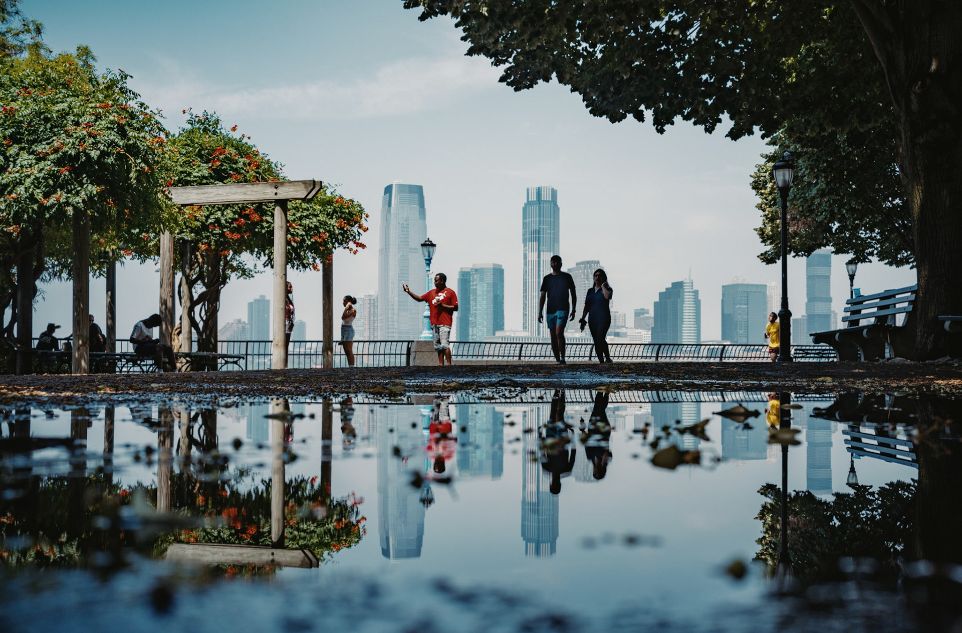captured in battery park, manhattan, after a summer rain, this scene gazes across the hudson river towards the soaring skyline of jersey city. the puddles mirror both the distant urban sprawl and the immediate, tranquil surroundings of the park, blending the vibrancy of new york with the calm of reflection. this moment, nestled between the city’s energy and a rare serenity, encapsulates the essence of manhattan’s waterfront charm.